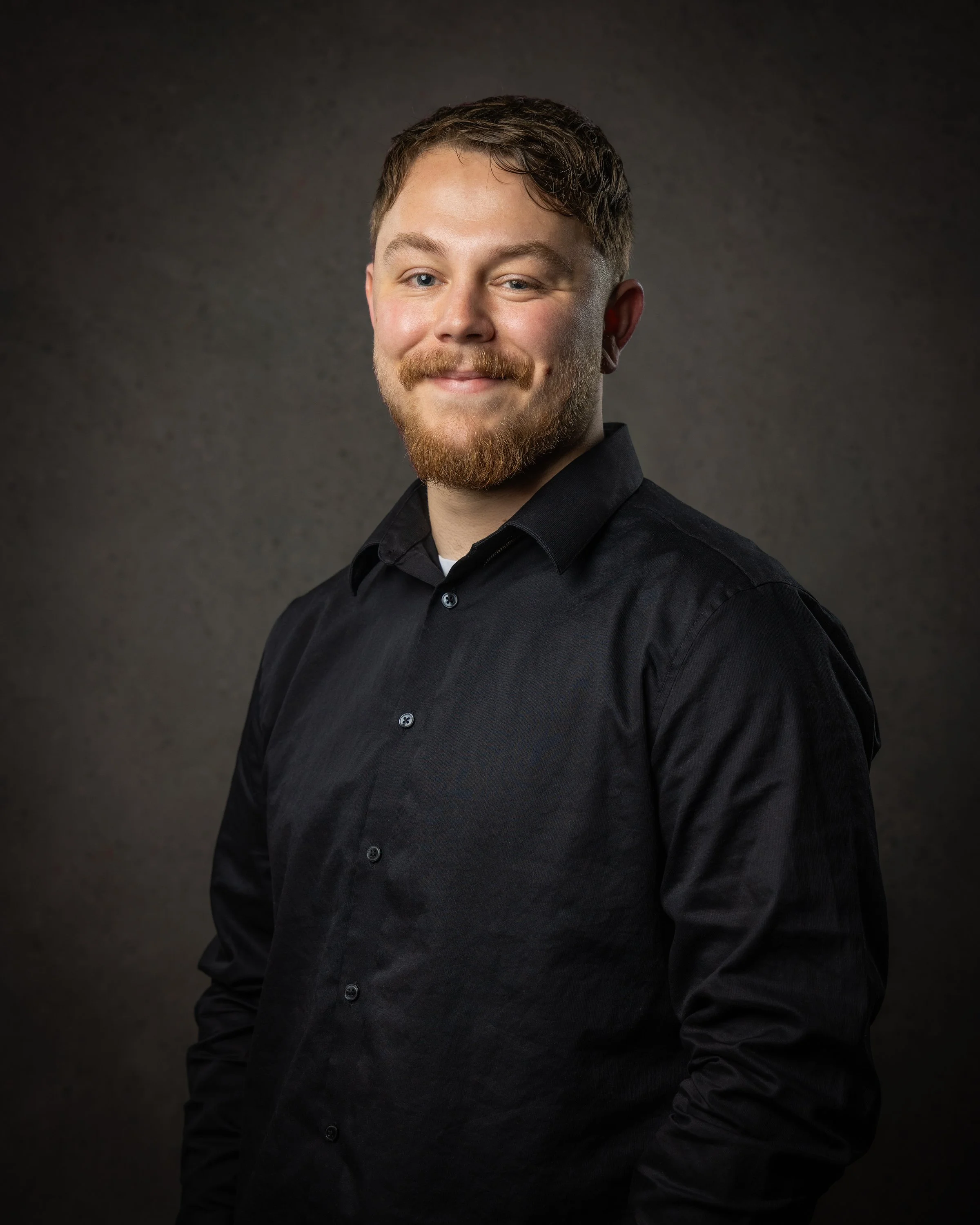 A young man with light skin, short curly brown hair, blue eyes, a mustache, and a beard, wearing a black shirt with a small colorful pattern. He is smiling slightly, head tilted to his right, in front of a dark textured background.