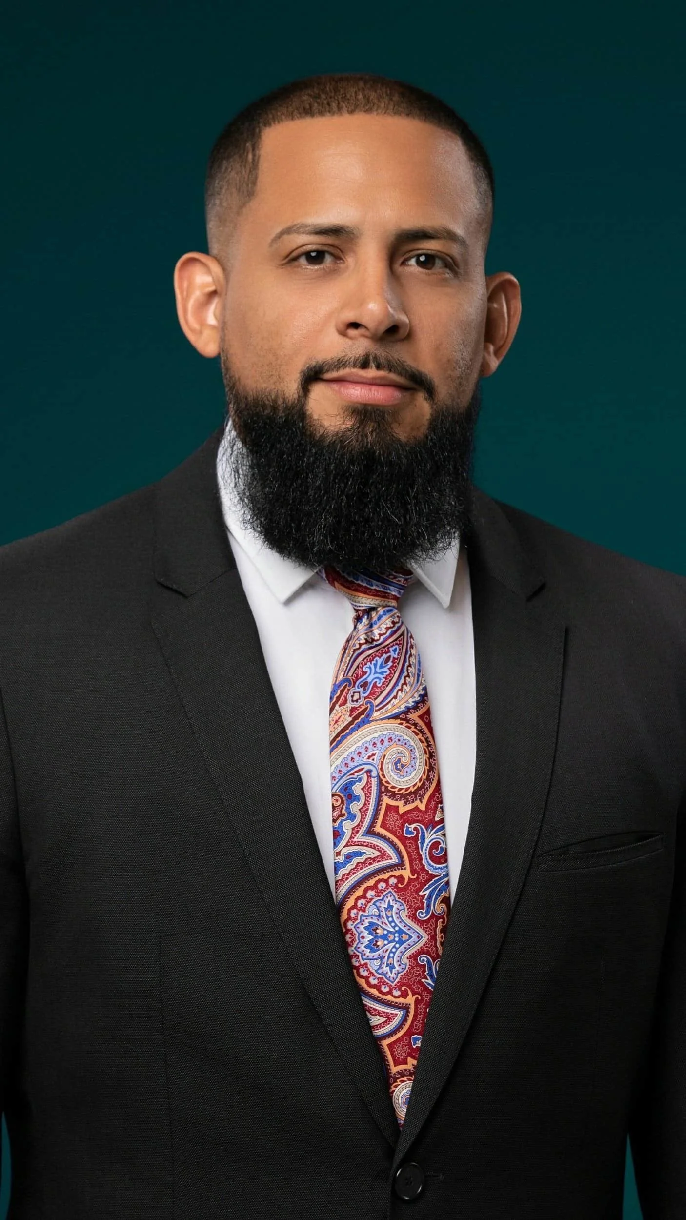 Man with a beard in a black suit and colorful paisley tie, standing in front of a dark teal background.