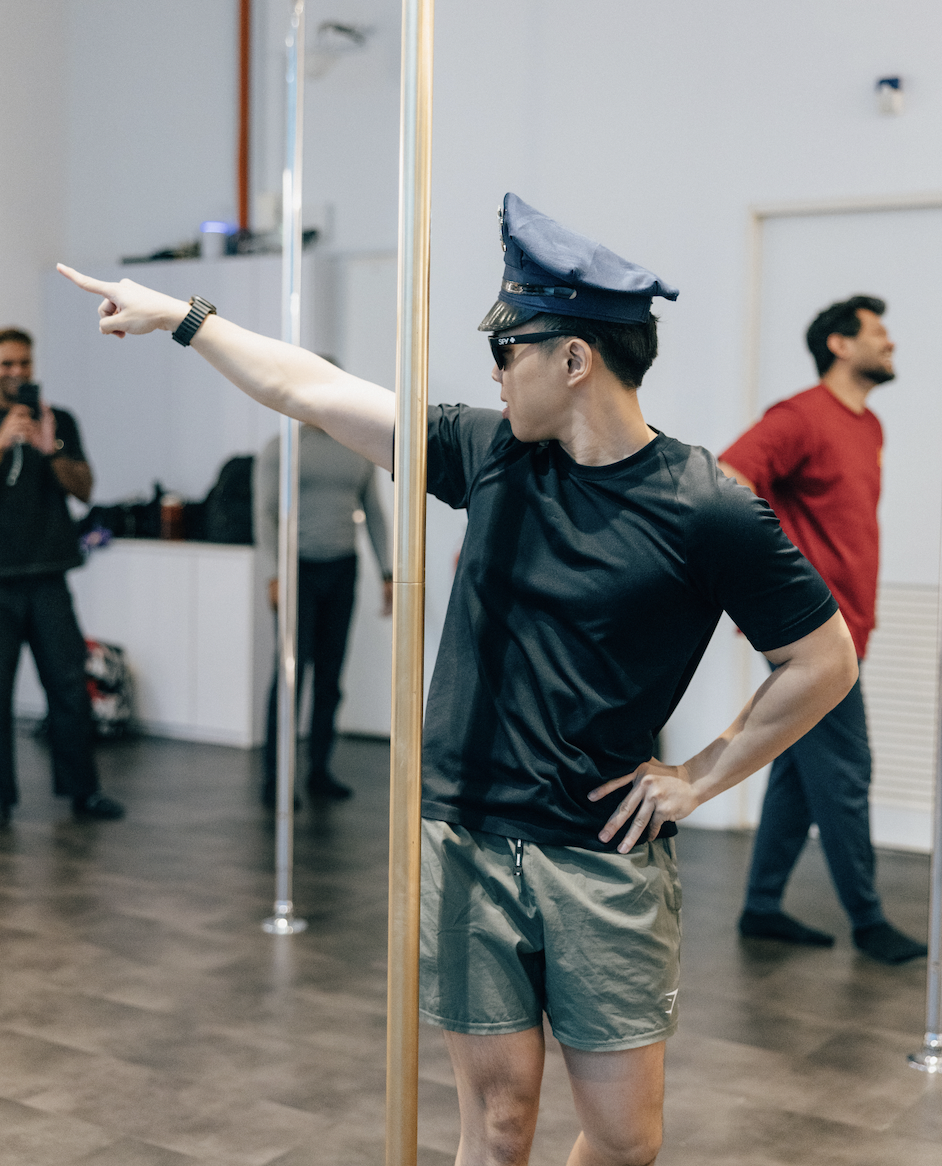 A person dressed as a police officer, wearing sunglasses and a police hat, pretending to point while standing next to a pole in a dance studio. Two men are visible in the background, one in a red shirt and the other taking a photo.