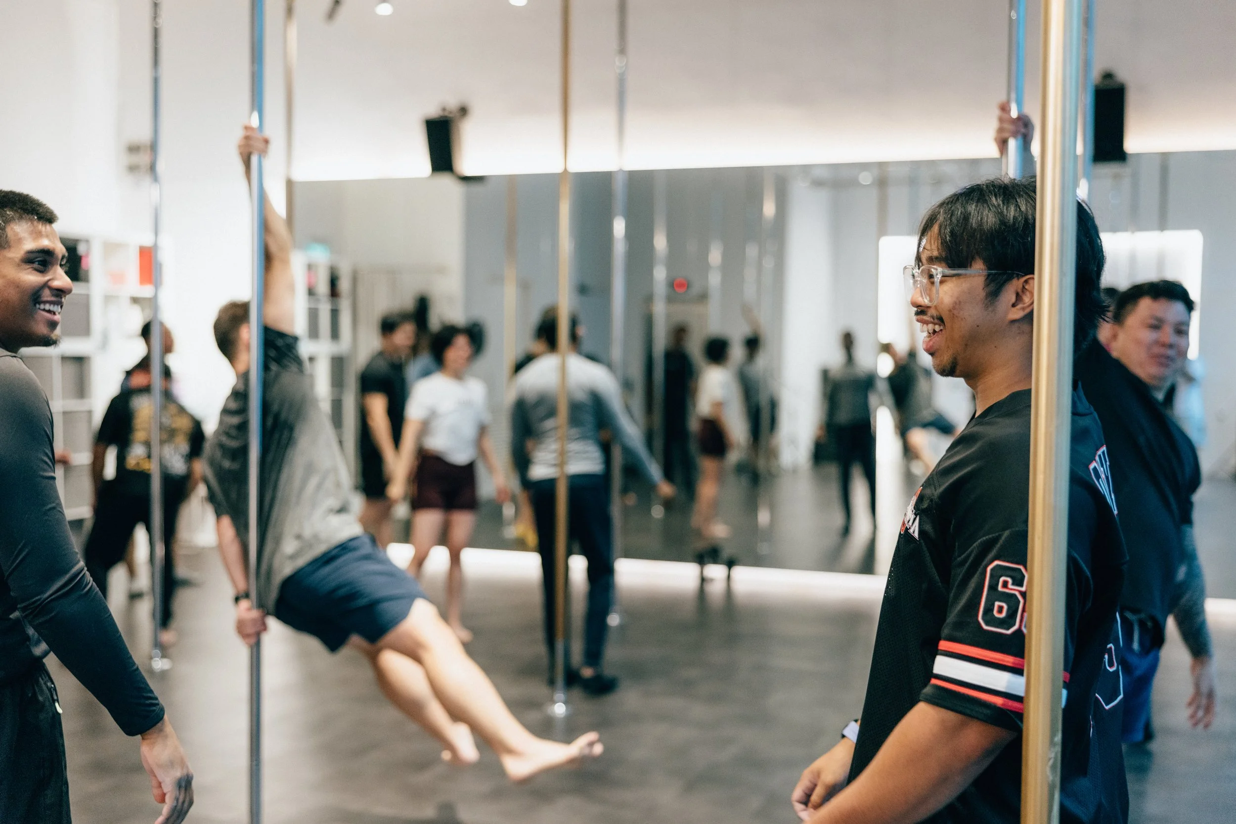 Two men are holding a pole between them while a woman performs a split in a dance studio. Several people are in the background, some watching and some walking around.