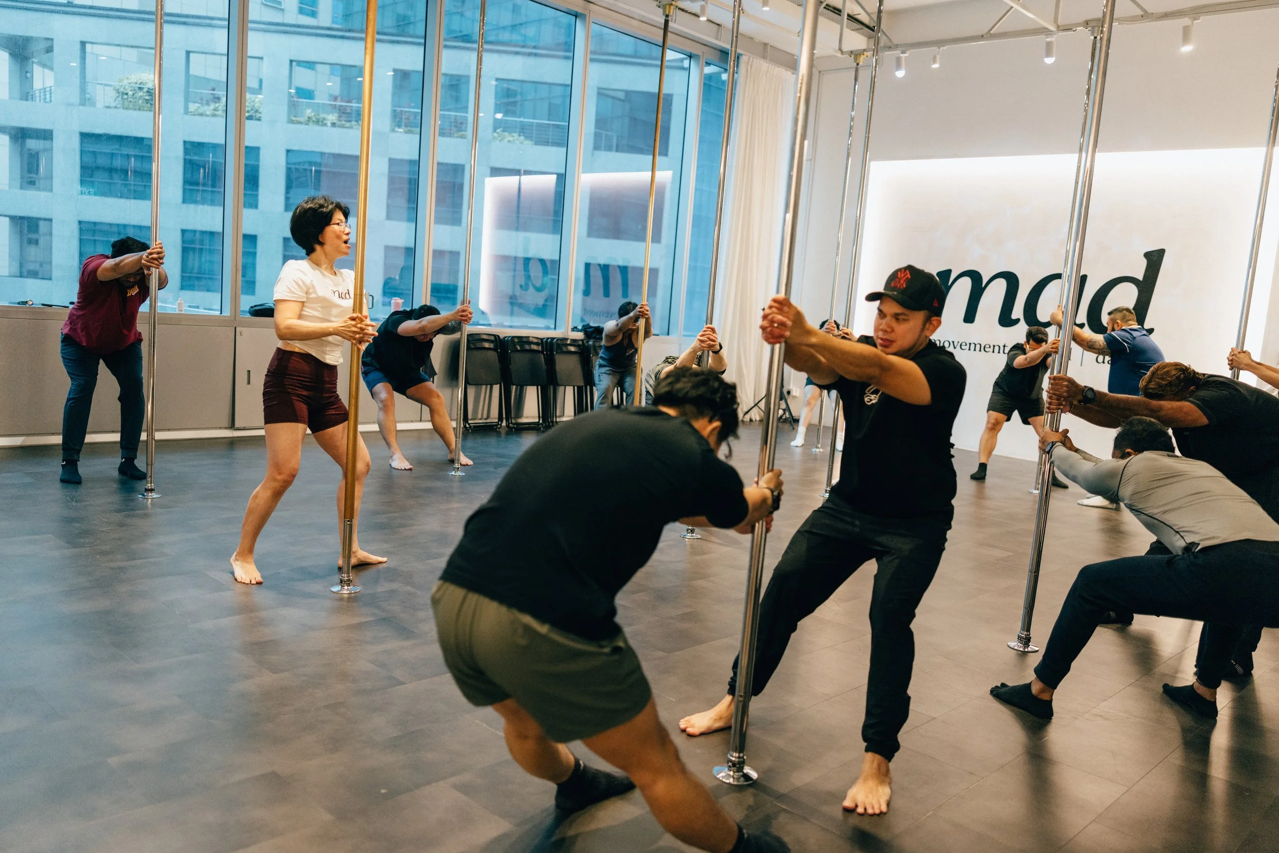 People in a dance studio practicing pole dancing, some are holding poles while stretching or balancing on the floor.