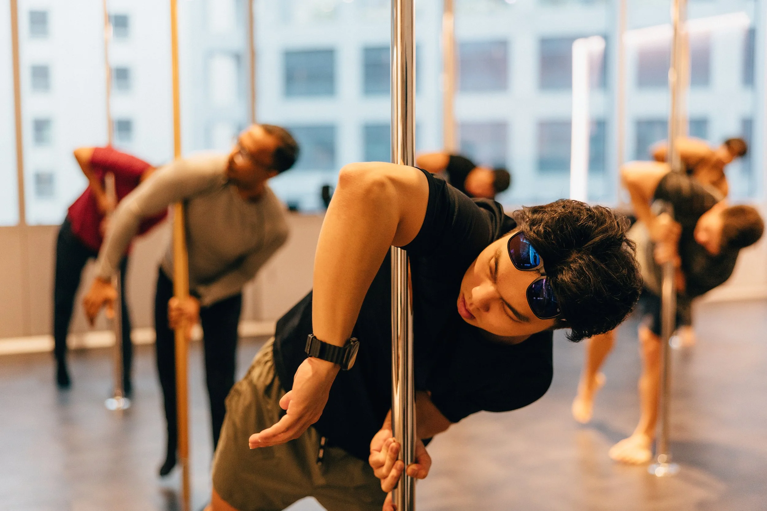 People practicing yoga in a studio, leaning on a vertical pole, with a city-view through large windows in the background.