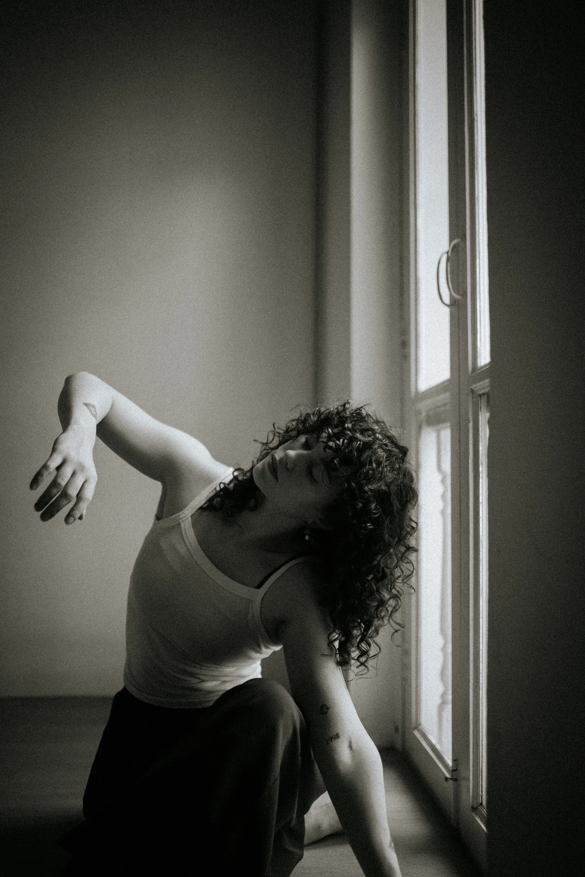 A woman with curly hair and tattoos on her arm is dancing or posing in front of a window with curtains, with natural light coming in from outside. She is wearing a tank top and appears to be in a minimalist interior space.