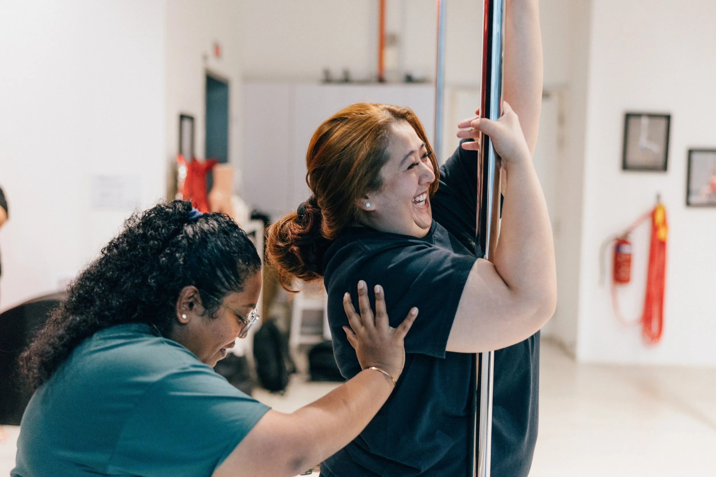 Women participating in a pole dancing class in a studio, smiling and supporting each other.