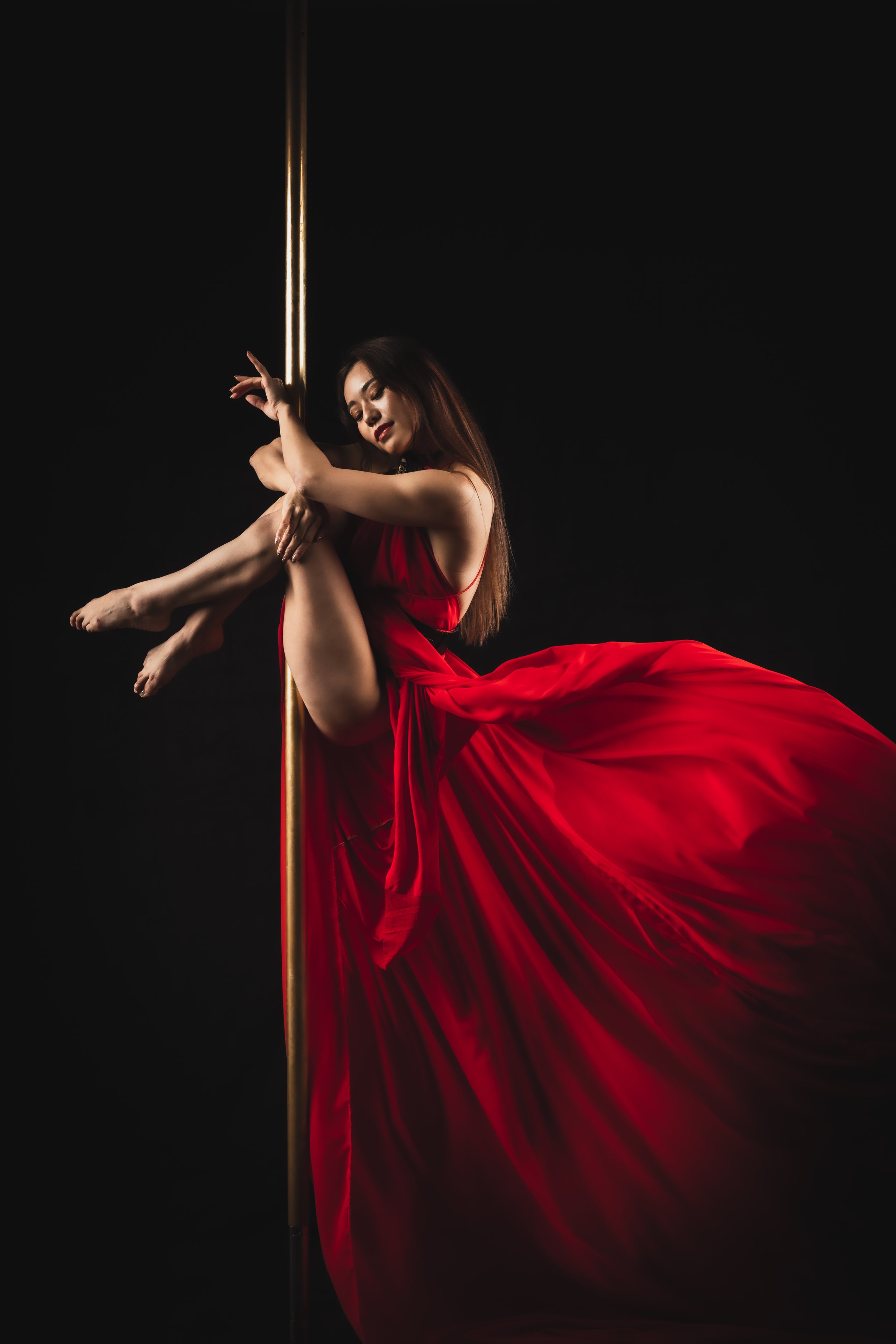 A woman in a red dress performing a pole dance against a black background, with dramatic lighting highlighting her and the pole.