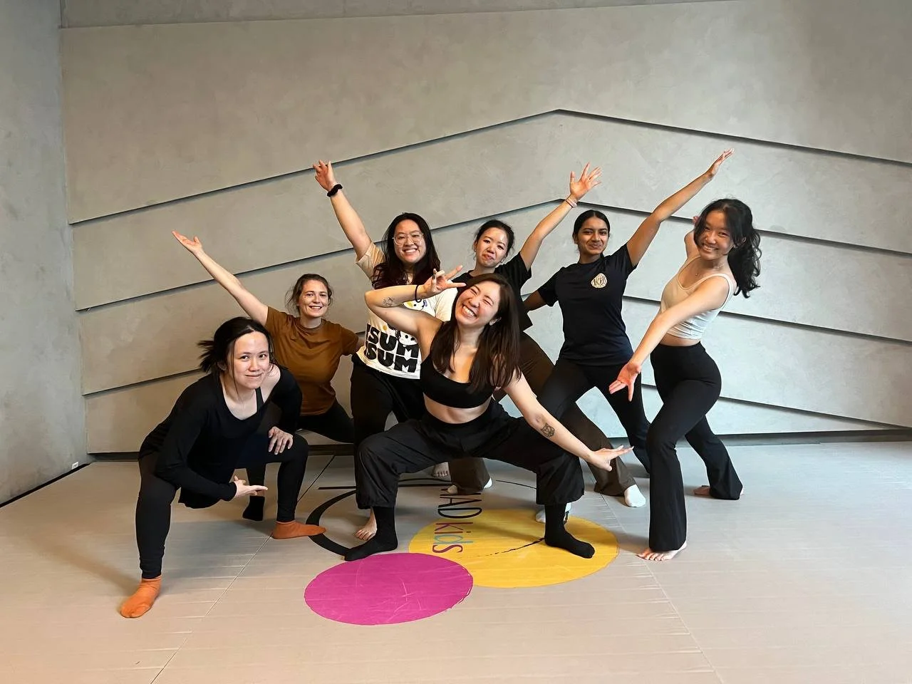 Seven women posing cheerfully in a dance studio with gray textured walls, some standing and others crouching or squatting, in various energetic and happy poses.