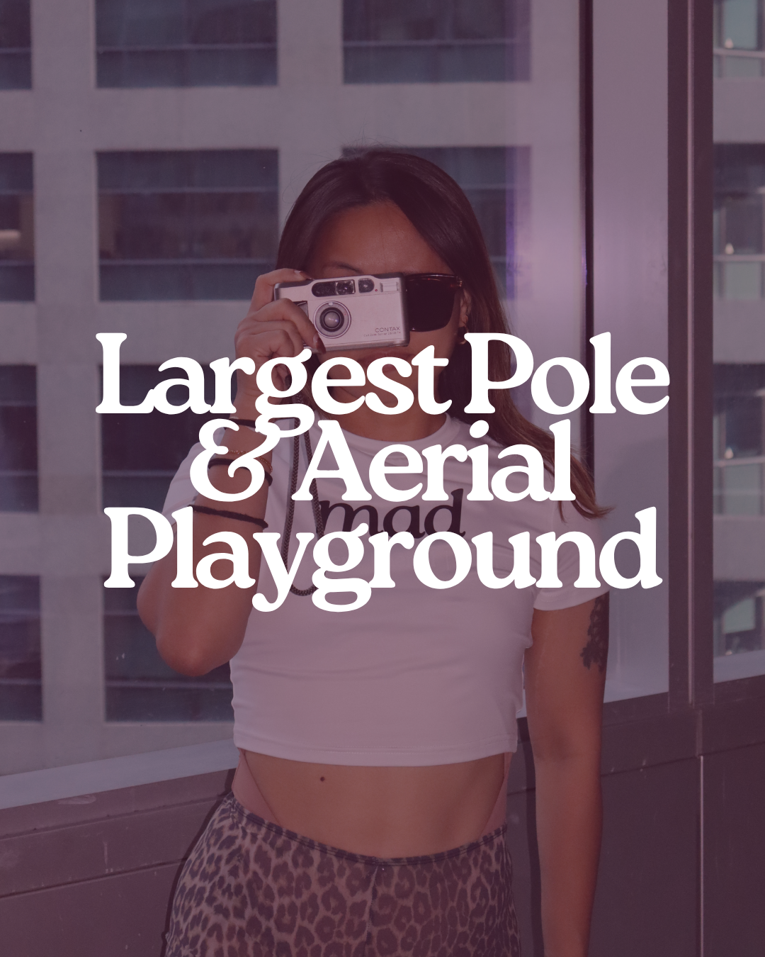 A woman with long dark hair taking a selfie with a camera while standing in front of a glass window of a tall building, with the text 'Largest Pole & Aerial Playground' overlaid.