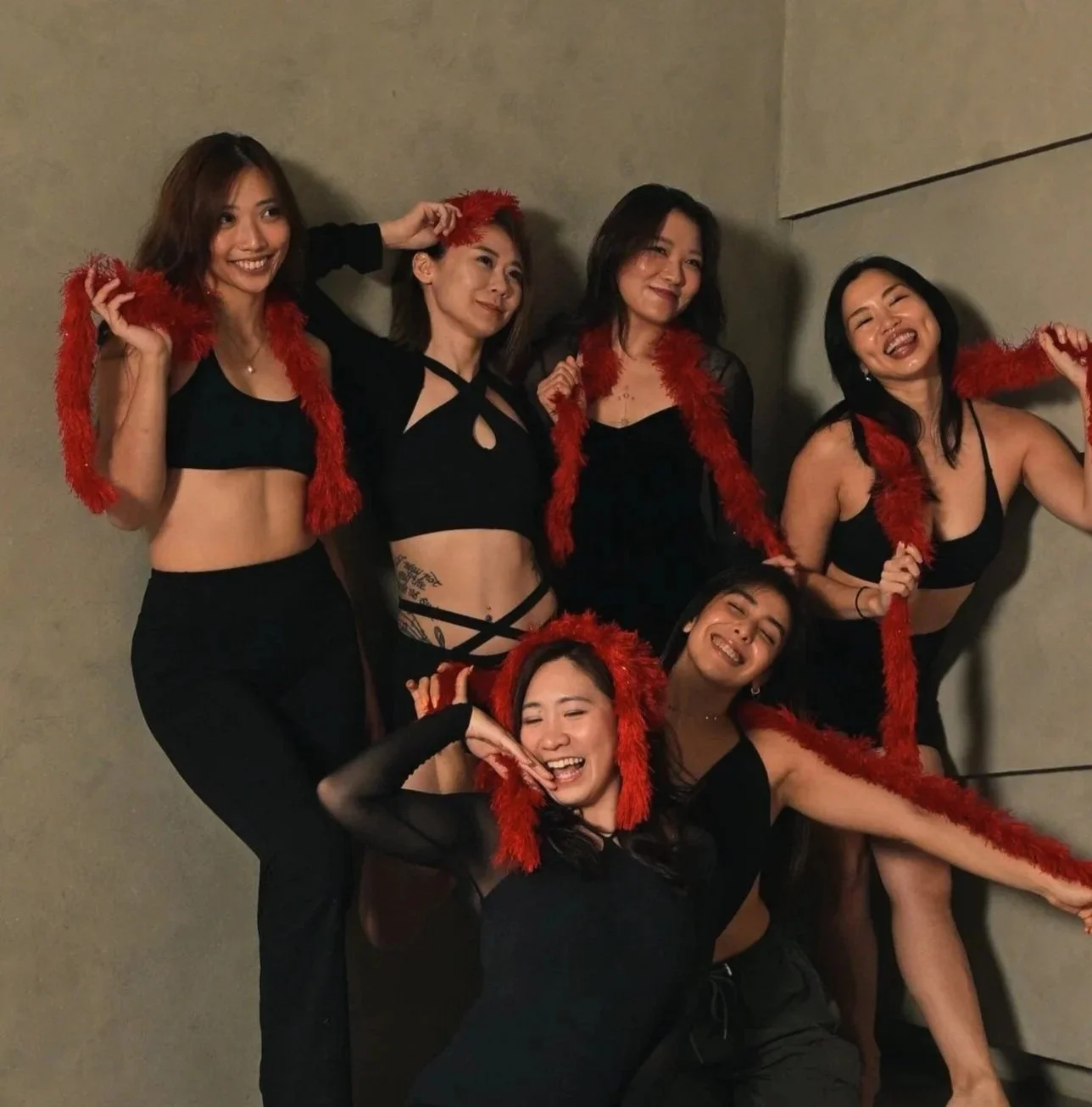 Group of six women posing together against a plain wall, wearing black outfits, red feather boas, and smiling happily.