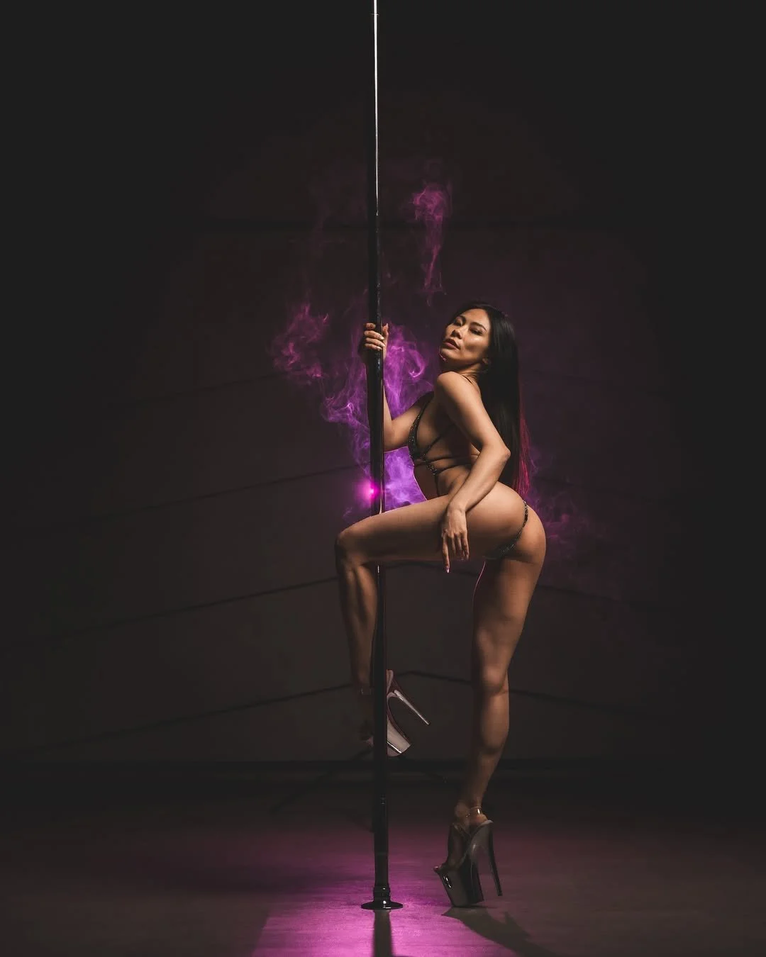 A woman in high heels and revealing lingerie poses with a pole against a dark background with purple smoke.