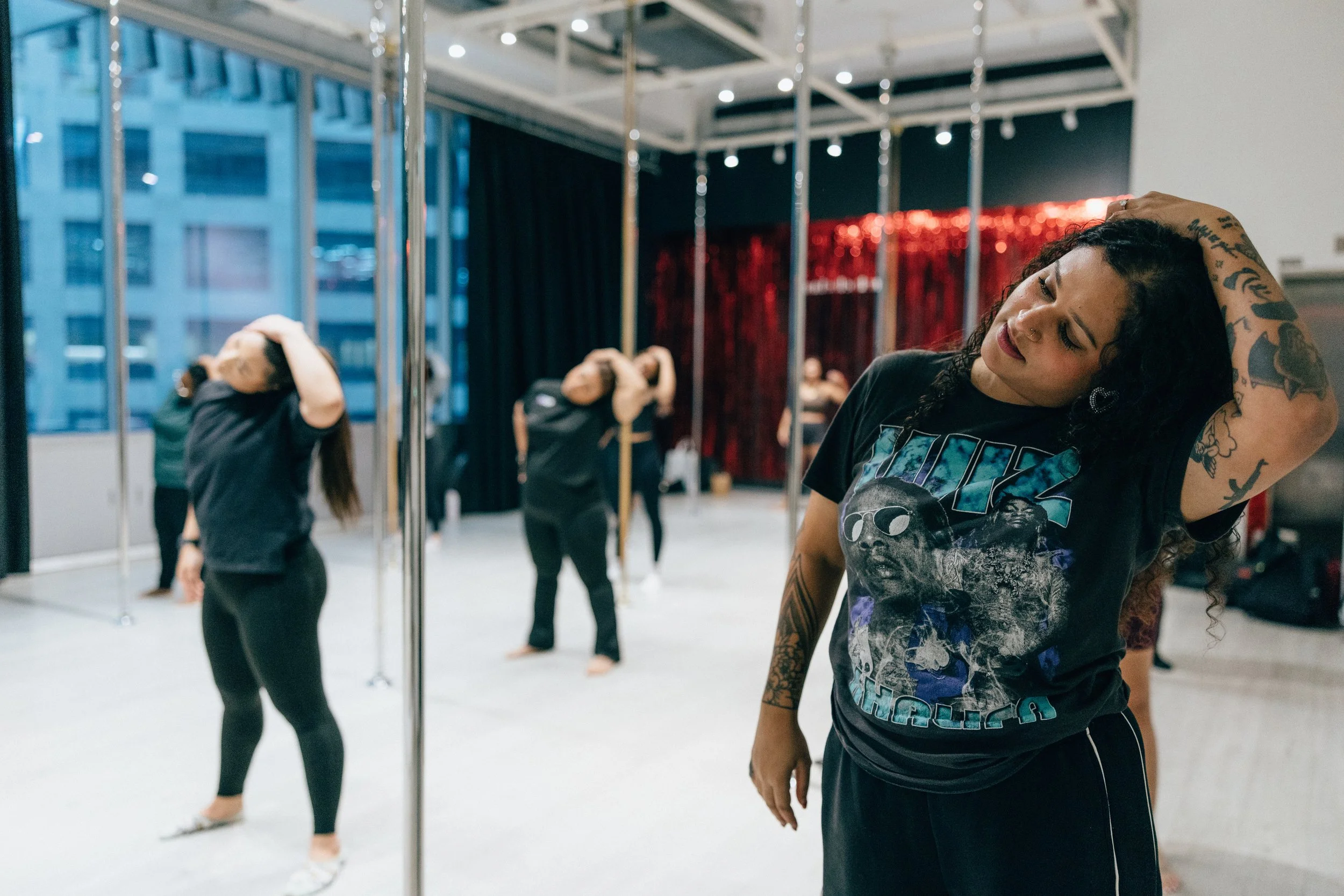A woman with tattoos and curly hair, wearing a graphic T-shirt, stands in a dance studio with a mirror, stretching her neck back. Other women are in the background, practicing dance moves, with studio poles and a cityscape visible through large windows.