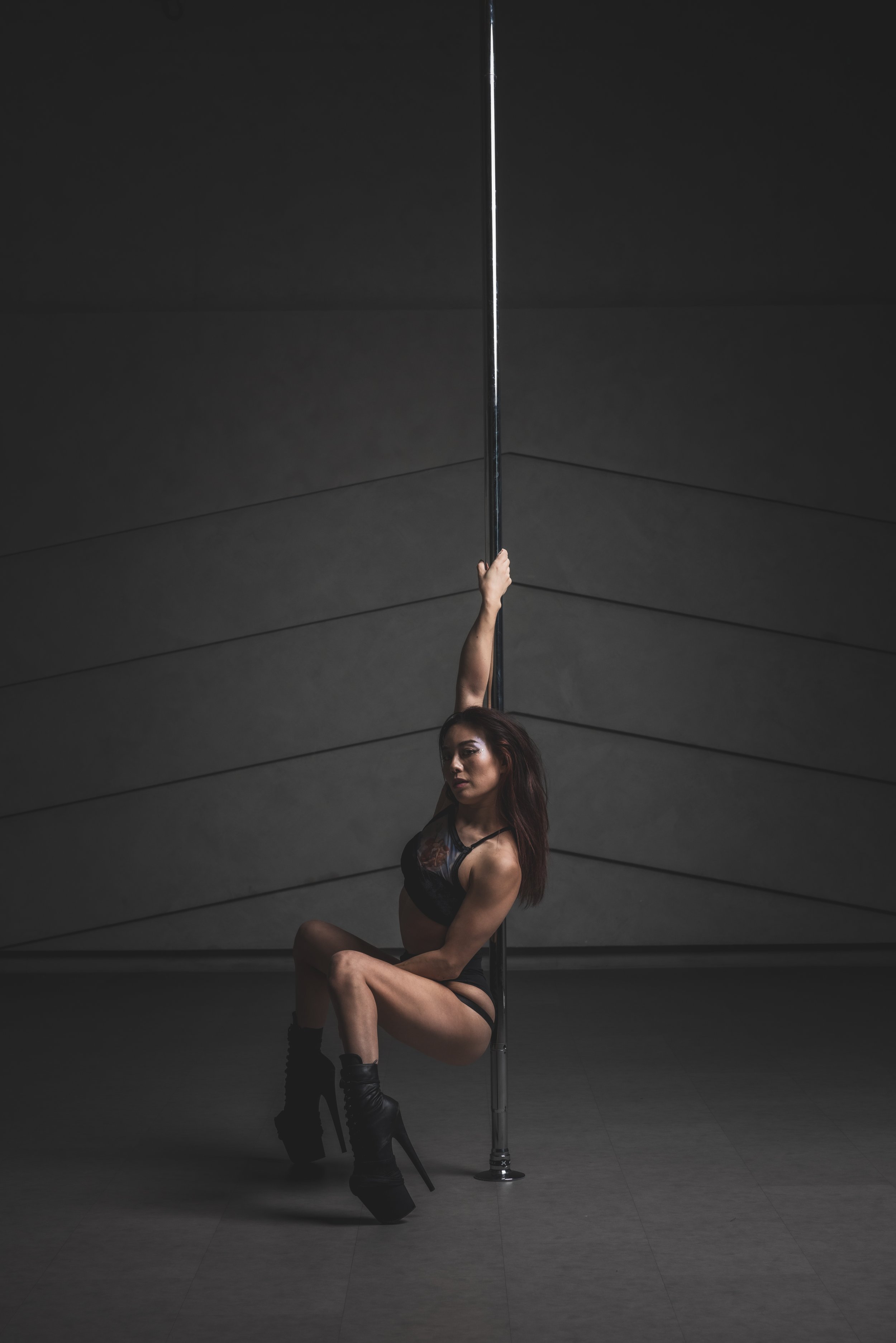 A woman with long red hair in black lace lingerie and high-heeled boots is sitting on the floor, holding a vertical pole with her left hand, in a dimly lit room with gray walls.