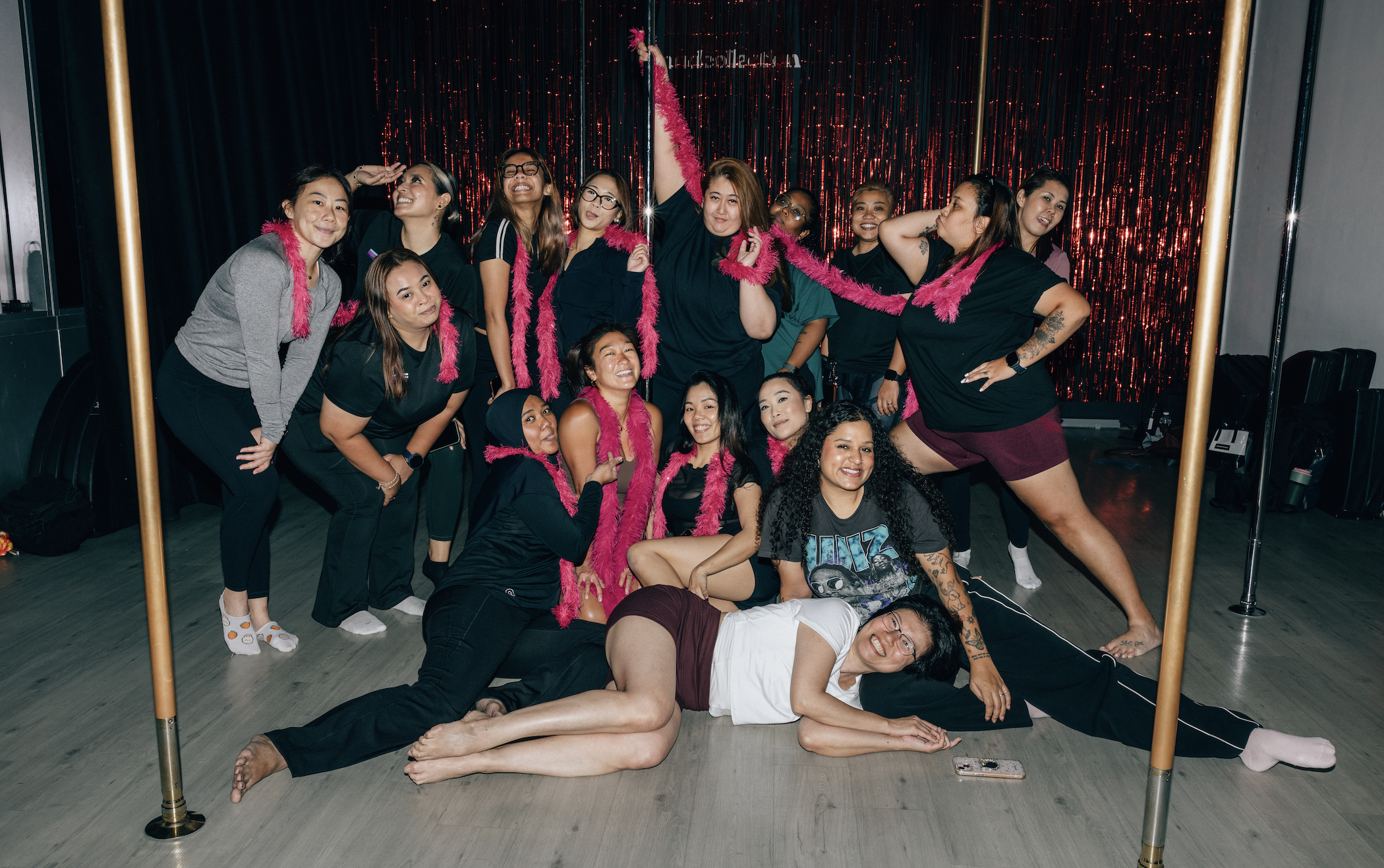 A group of women posing together at a social event, some standing and some sitting or lying on the floor, with a black and red glitter backdrop, some wearing pink feather boas, and a person holding a pink feather duster.