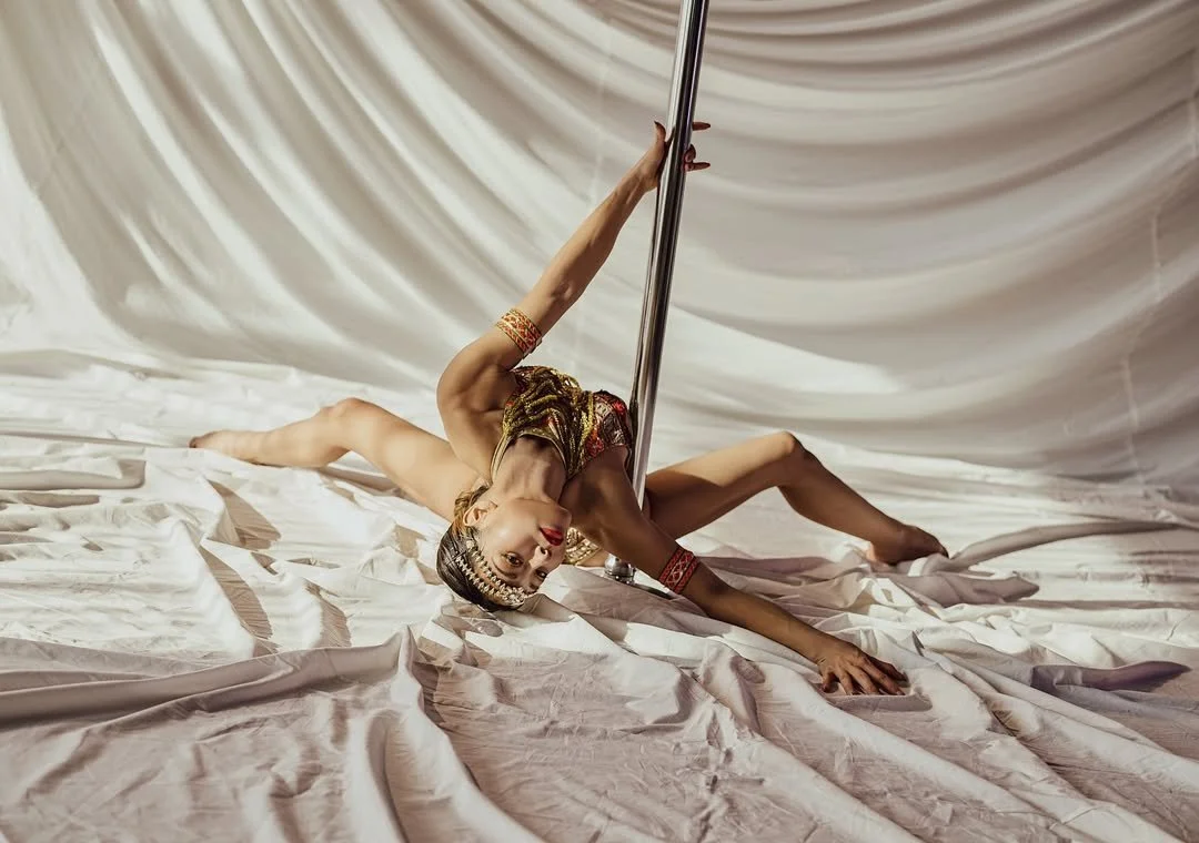 A female pole dancer in a colorful costume performing on a stage with white curtains, lying on the floor and holding a vertical pole.