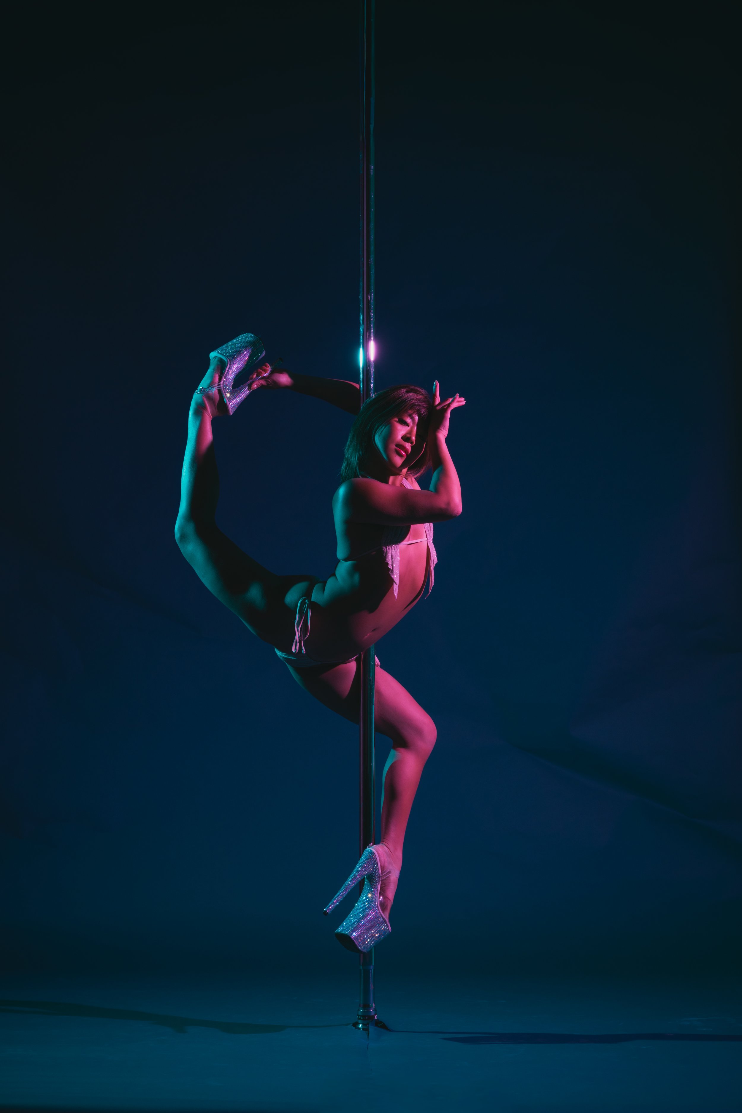 A female pole dancer performing a pose upside down on a vertical pole, wearing sparkly high heels and a pink outfit, with a dark background and colorful lighting.