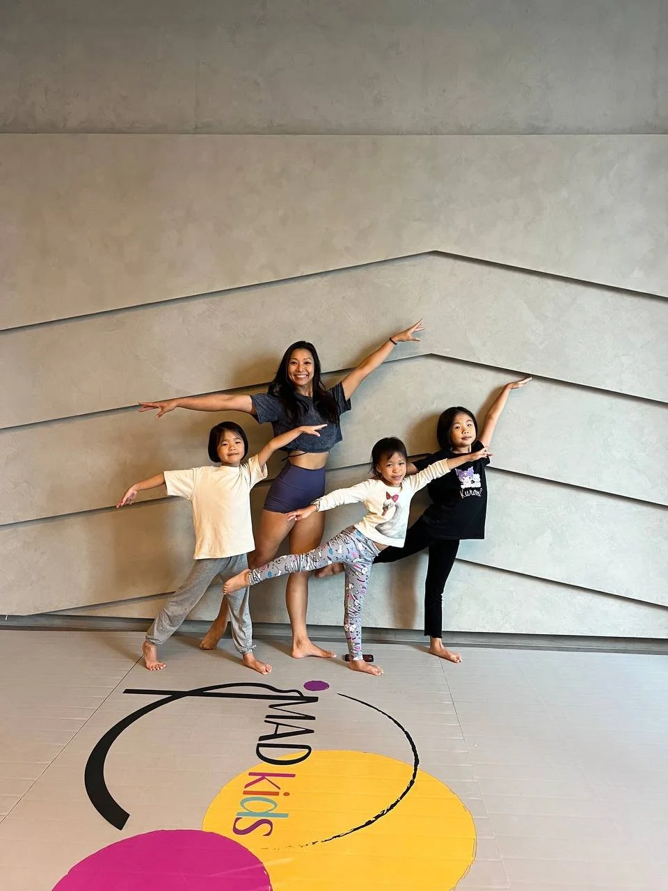 Five people, one adult woman and four children, practicing ballet or dance poses against a textured gray wall. They are standing on a colorful floor with the words 'STUDIO Kids' written on it.