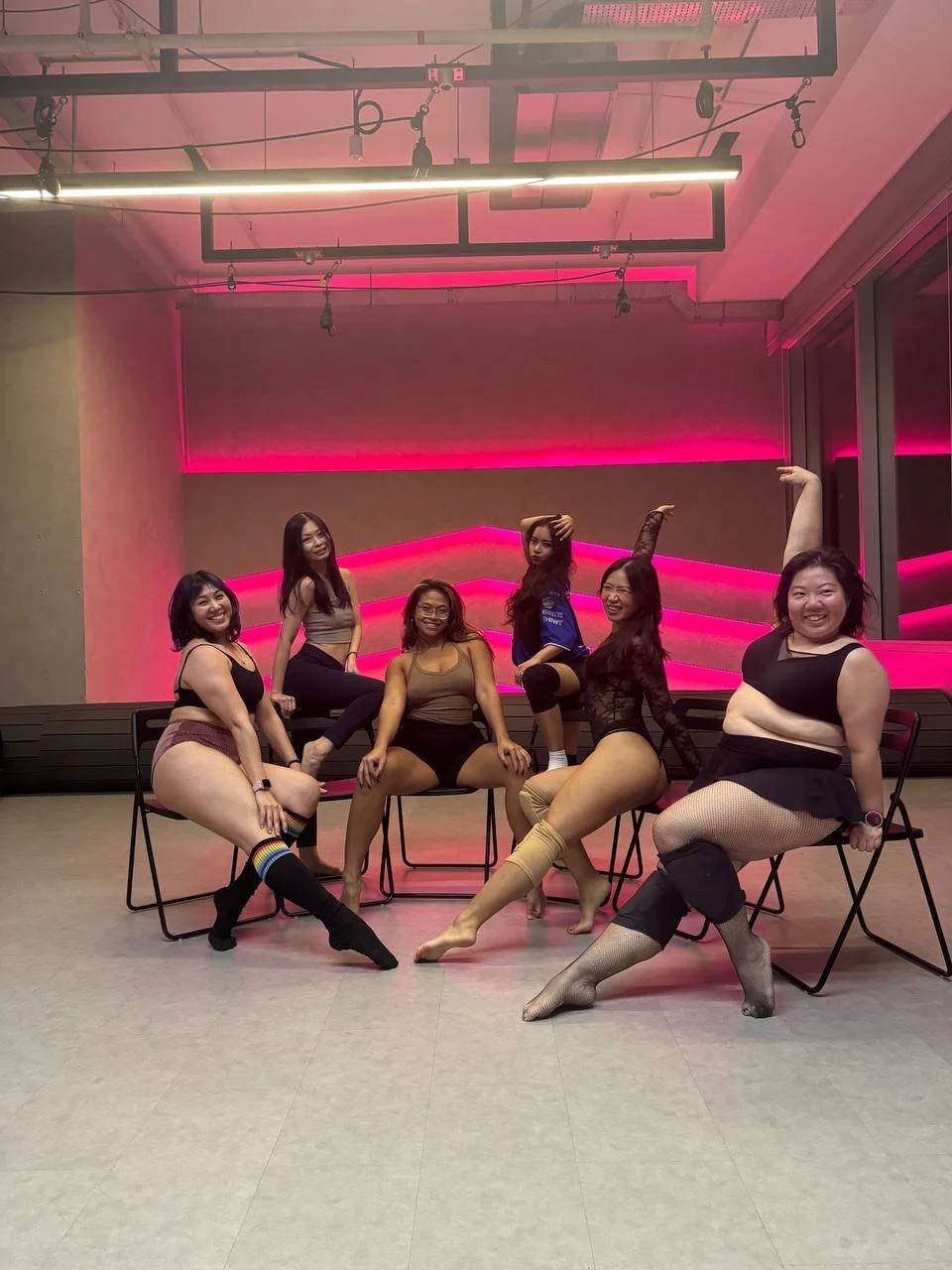 Five women posing in a room with pink neon lighting, sitting on chairs and in various dance or yoga poses, wearing athletic or dancewear including fishnet stockings and colorful striped socks.