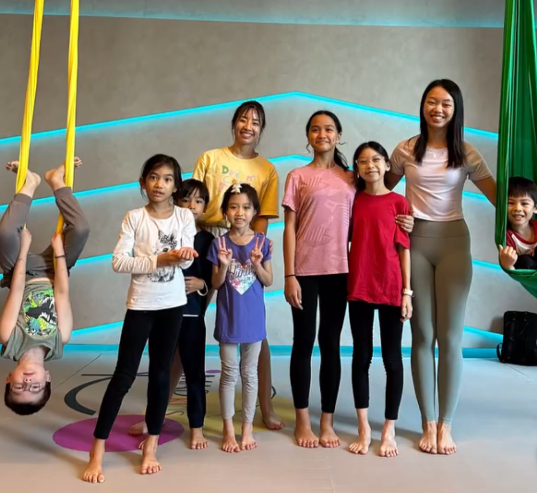 Group of children and two adult women smiling indoors, some children holding colorful aerial silk fabrics.