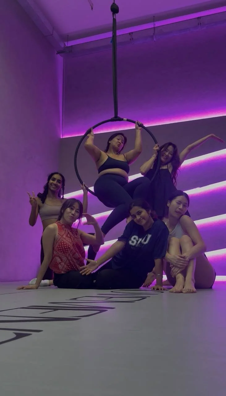 Group of five women posing together in a dance or performance studio with purple neon lighting, one woman sitting in a hoop hanging from the ceiling, others surrounding her in various poses.