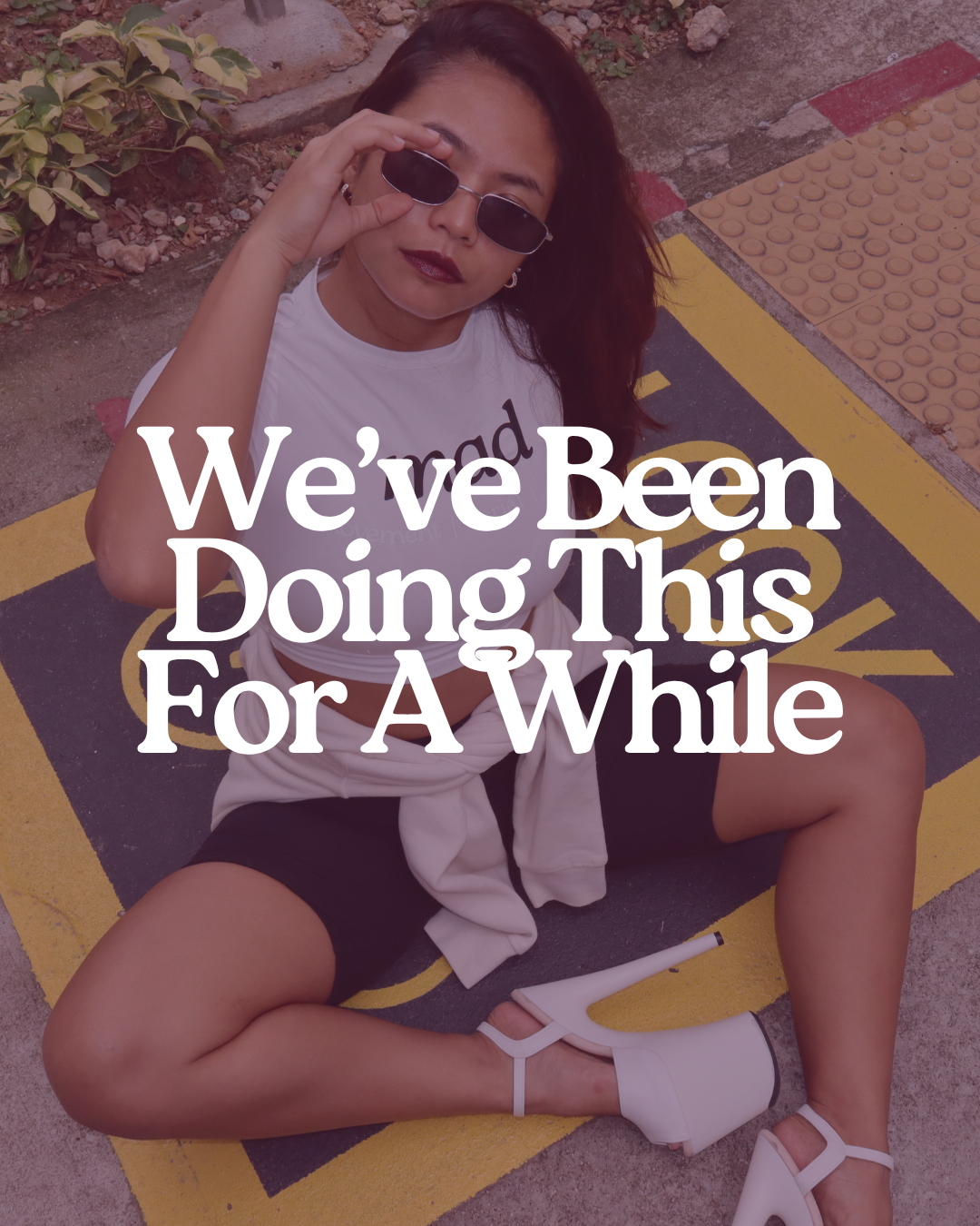 A woman with dark hair and sunglasses sitting on a yellow and black parking lot marking, wearing a white sleeveless top, black shorts, white high heels, and a white sweatshirt tied around her waist, with a slight smile and holding her sunglasses