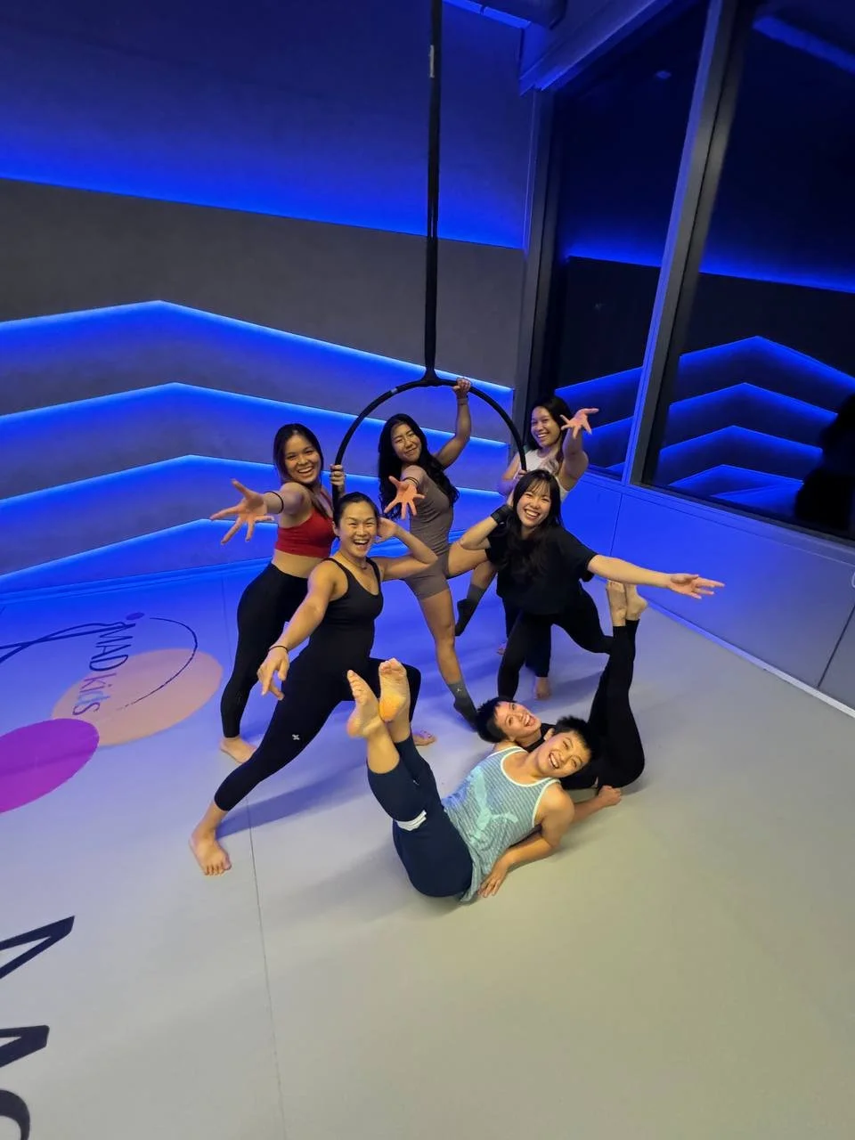 Six women in athletic wear posing cheerfully inside a dance or fitness studio with blue lighting, one woman doing a split on the floor, others standing around her with extended arms, and one hanging from a circular aerial hoop.