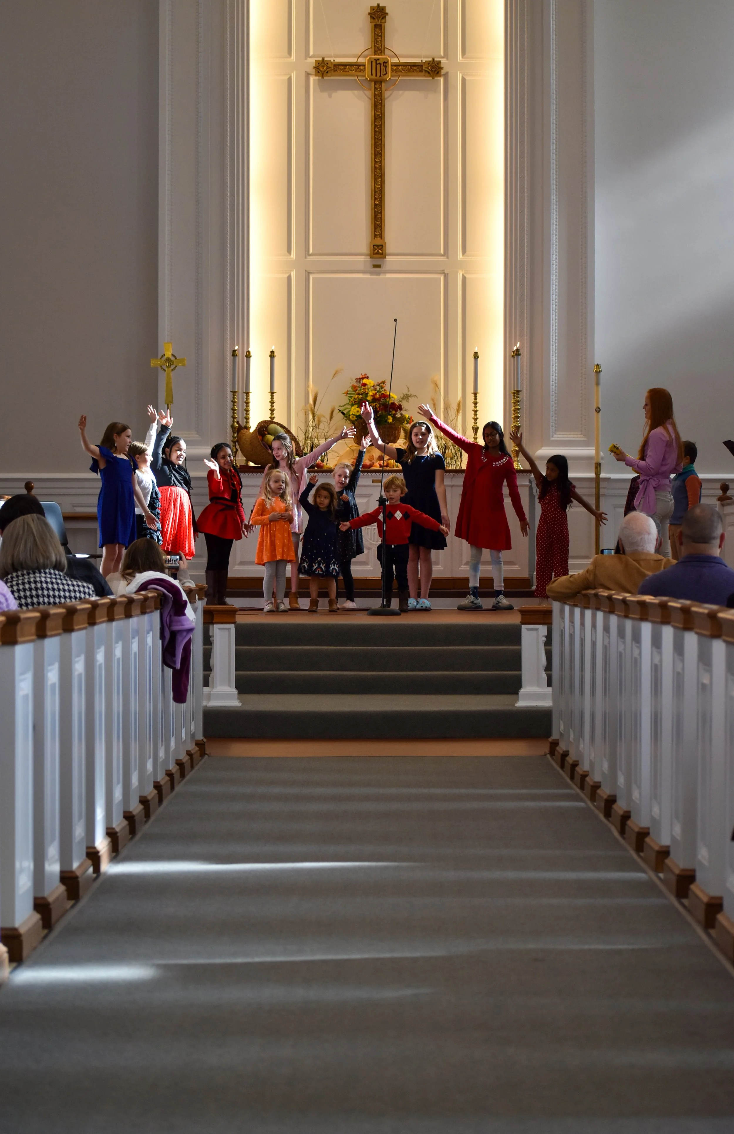 The children's choir performs in the sanctuary, with arms raised, in front of a large cross