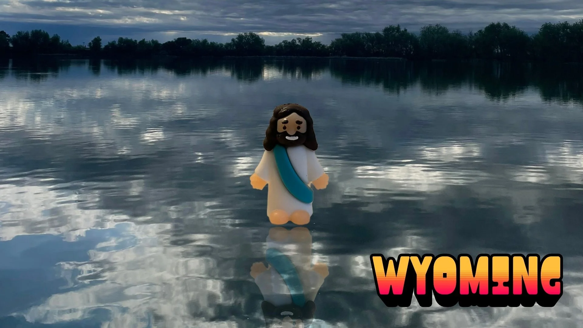Tiny Jesus figurine standing on a calm lake at dusk, trees silhouetted on the horizon. Text reads WYOMING.