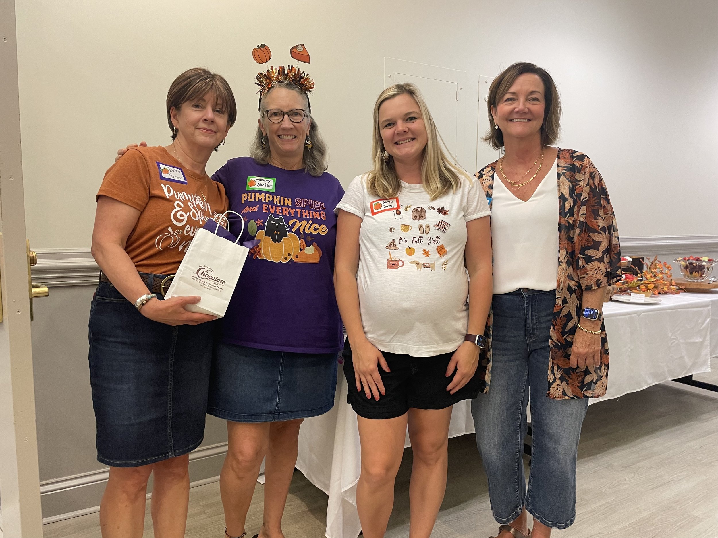 Four women standing together at an indoor gathering, smiling at the camera, with one wearing a pumpkin-themed headband, and they are dressed casually.