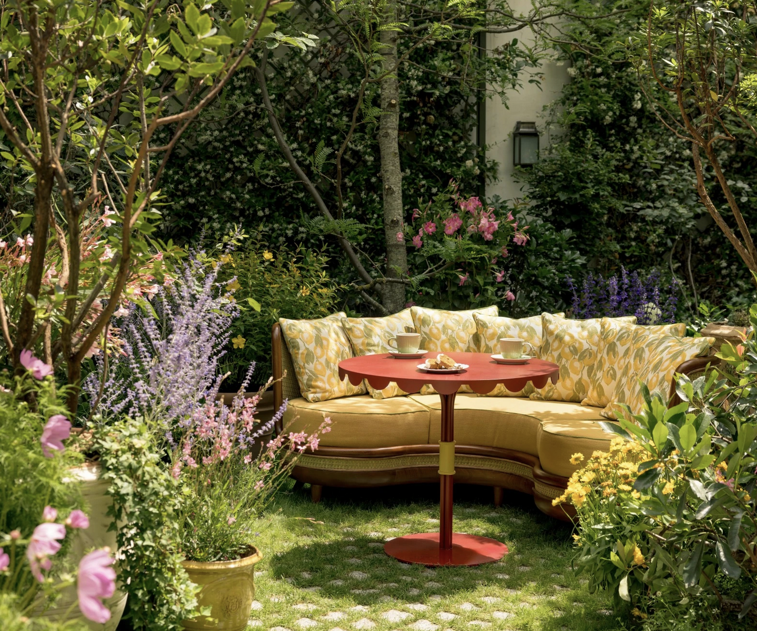 A garden scene with a yellow outdoor sofa with patterned cushions, a red round table with tea cups and snacks, and lush green plants and colorful flowers surrounding the area.