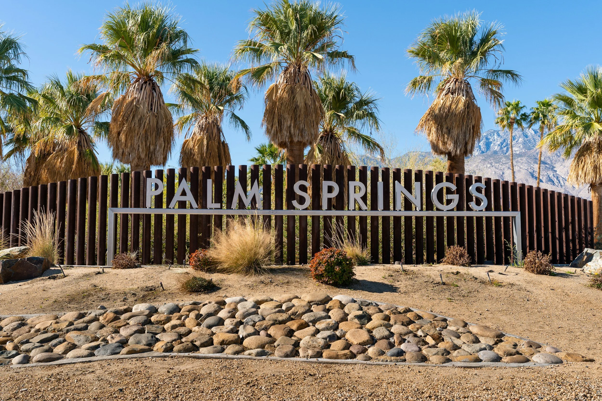 Entrance sign for Palm Springs with tall palm trees, desert plants, and dry landscape under a clear blue sky. Mountain range in the background.