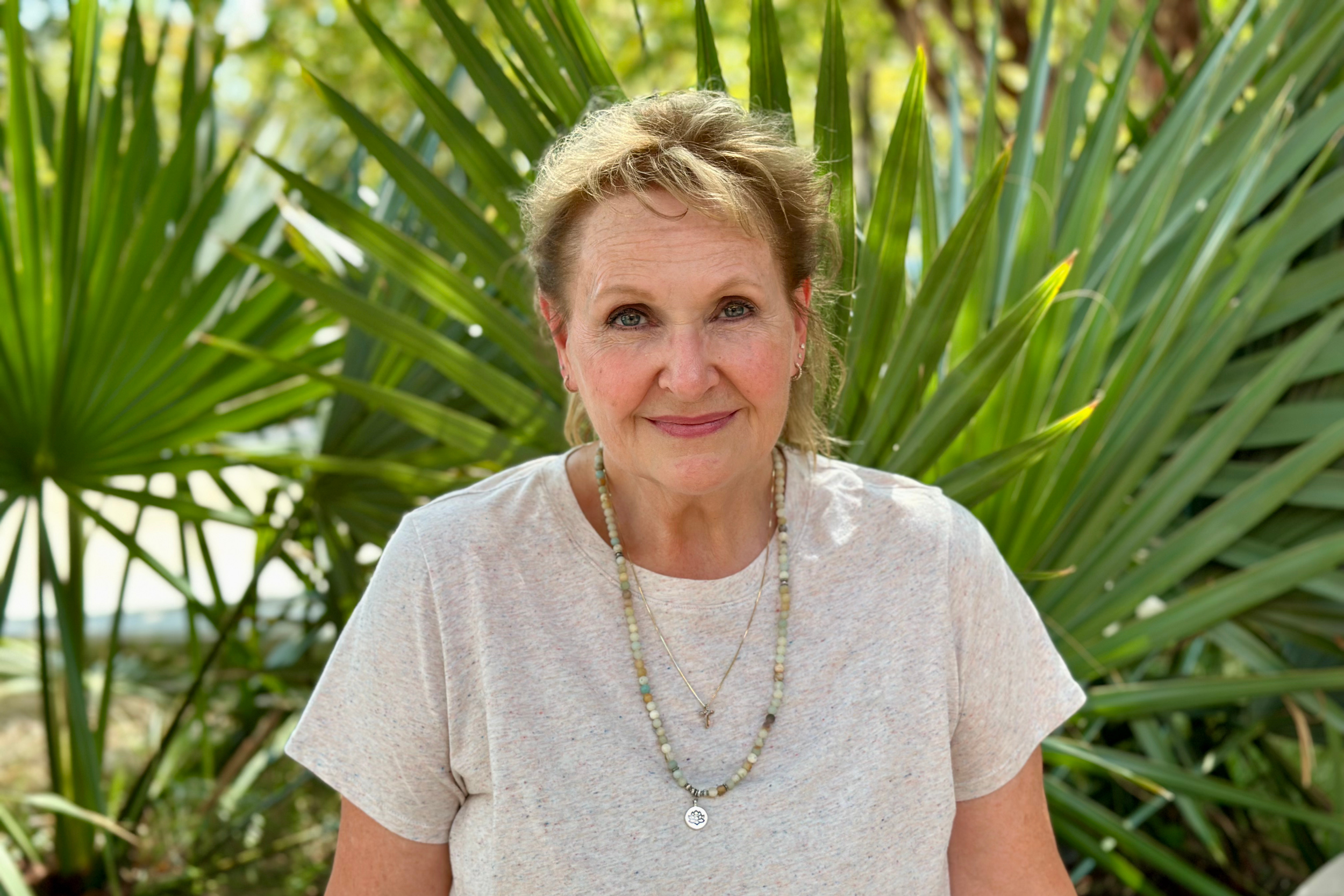 A blonde woman wearing an ivory tee, smiling warmly in front of palm trees.