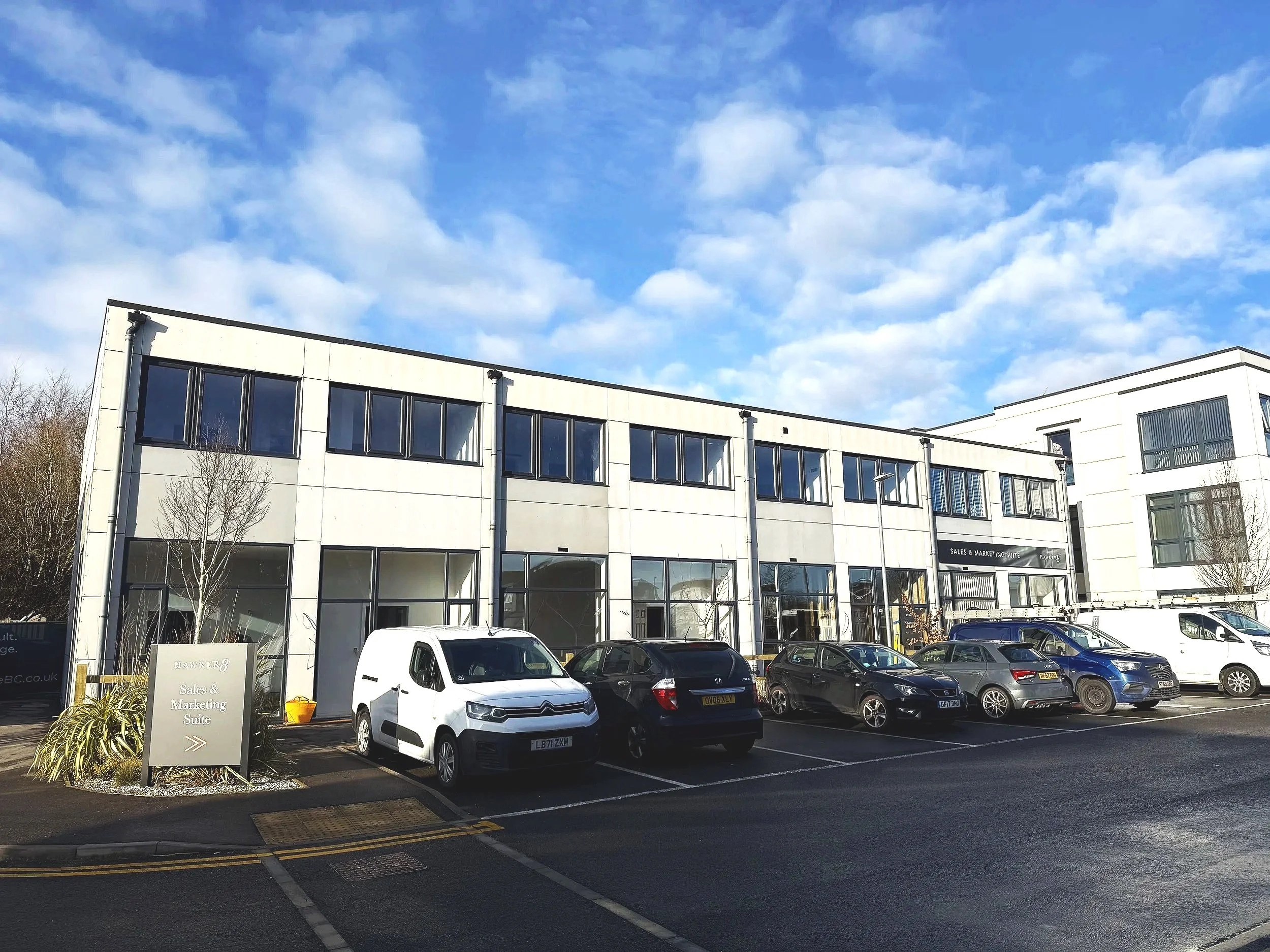 Commercial office building with parking lot and several cars in front under a partly cloudy sky.