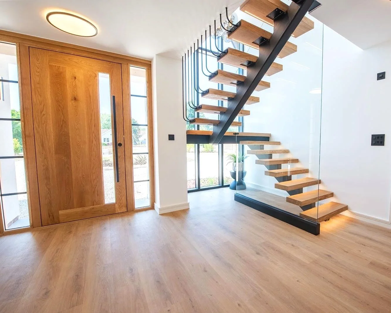 Interior view of a modern house with wooden front door, large windows, and floating wooden staircase with black metal supports and glass railing.