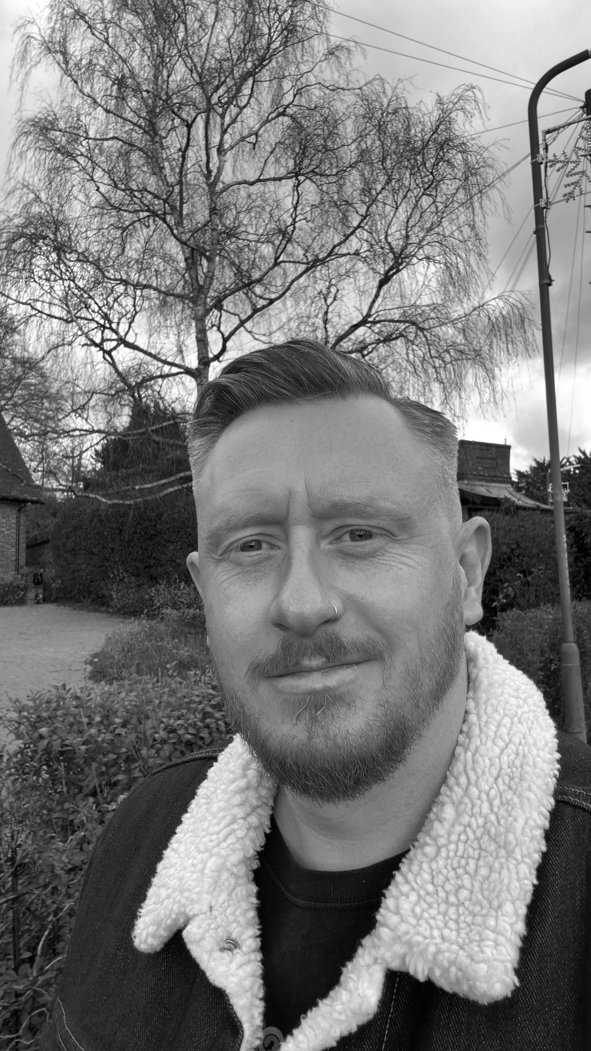 A black and white photo of a man with a nose piercing and a beard, smiling slightly outdoors with trees, bushes, and a streetlight in the background.