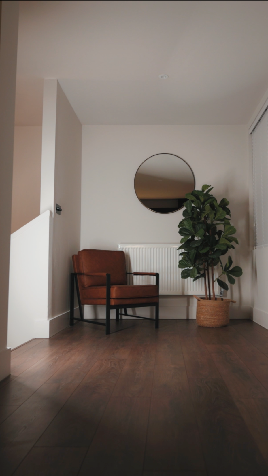 A cozy interior corner featuring a brown leather armchair with black metal legs, a large green potted plant in a woven basket, a white radiator, a round mirror on the wall, and hardwood flooring.