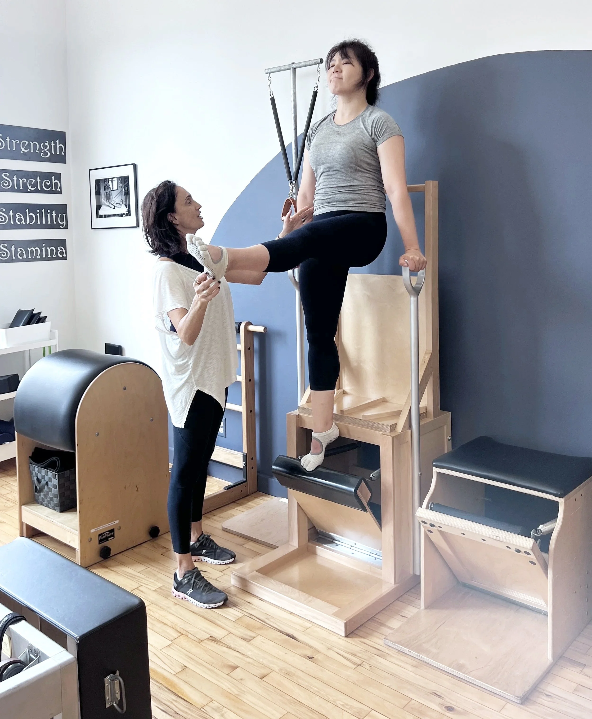 A woman is doing Dips on the High Chair with assistance from a trainer in a Pilates studio.