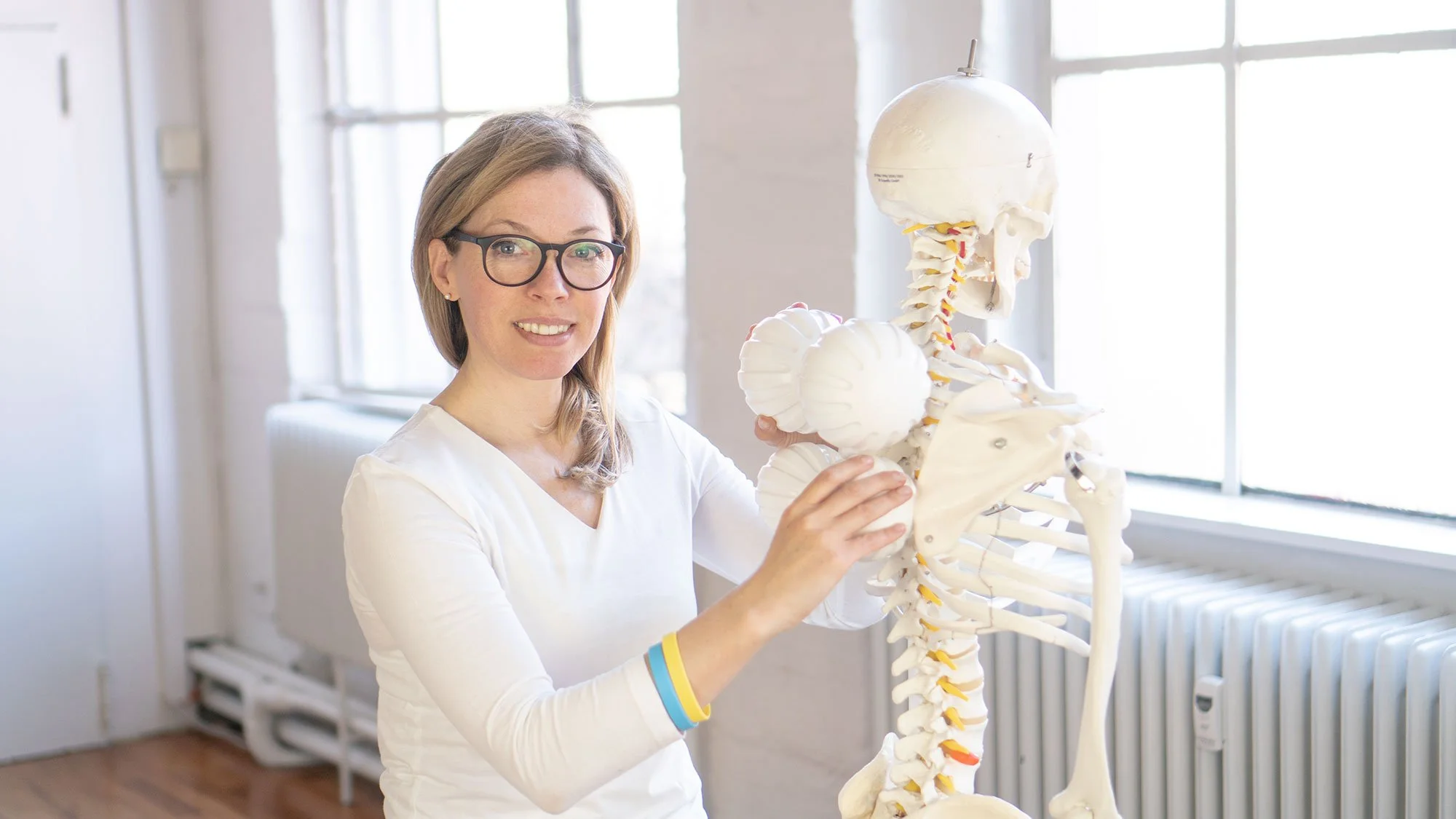 A woman wearing glasses and a white shirt demonstrates a human spine model in a well-lit room.