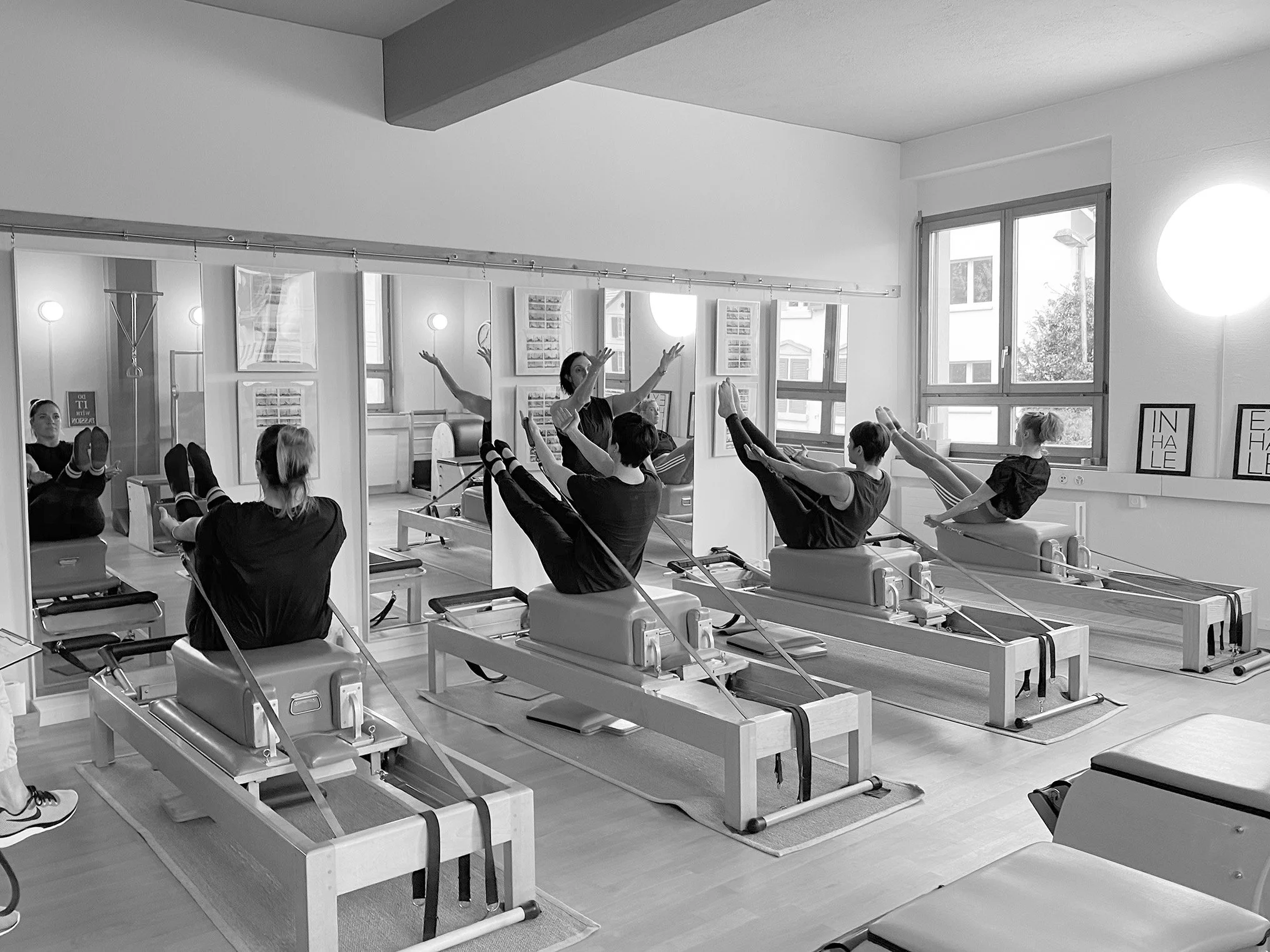 A group of people participating in a Pilates  class using reformer machines, guided by an instructor in a well-lit studio with large windows.