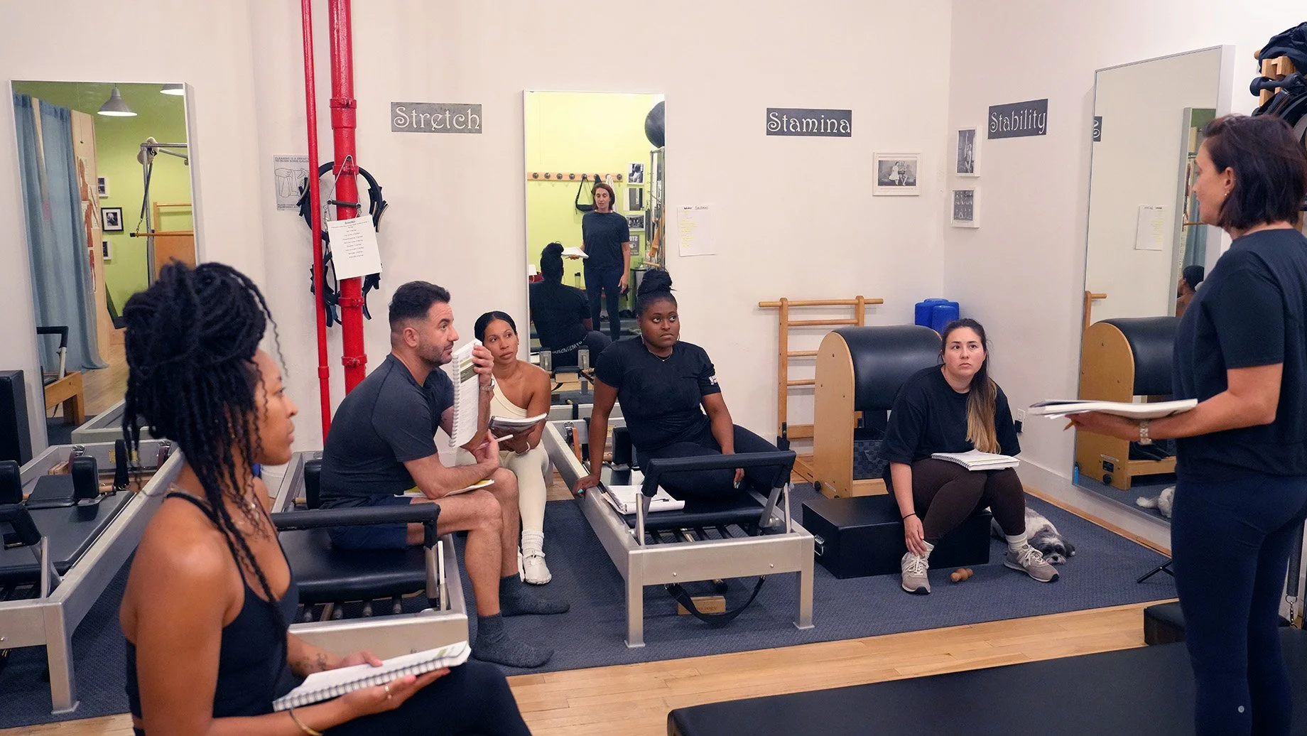 Group of people in a Pilates studio listening to instructor, some seated on chairs, with workout equipment and mirrors on the walls.