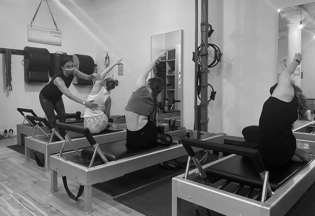 Four women participating in a Pilates class inside a studio. One instructor helps two women with their exercises, while a third participant performs a stretch on her reformer machine. All are on reformer Pilates equipment.