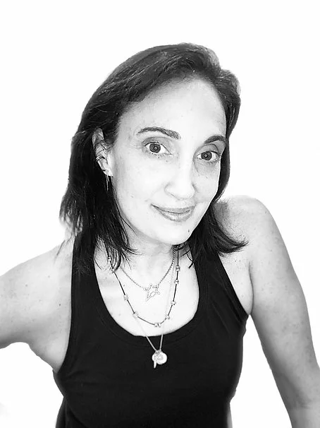Black and white photo of a woman with shoulder-length dark hair, wearing a black sleeveless top and layered necklaces, smiling at the camera.