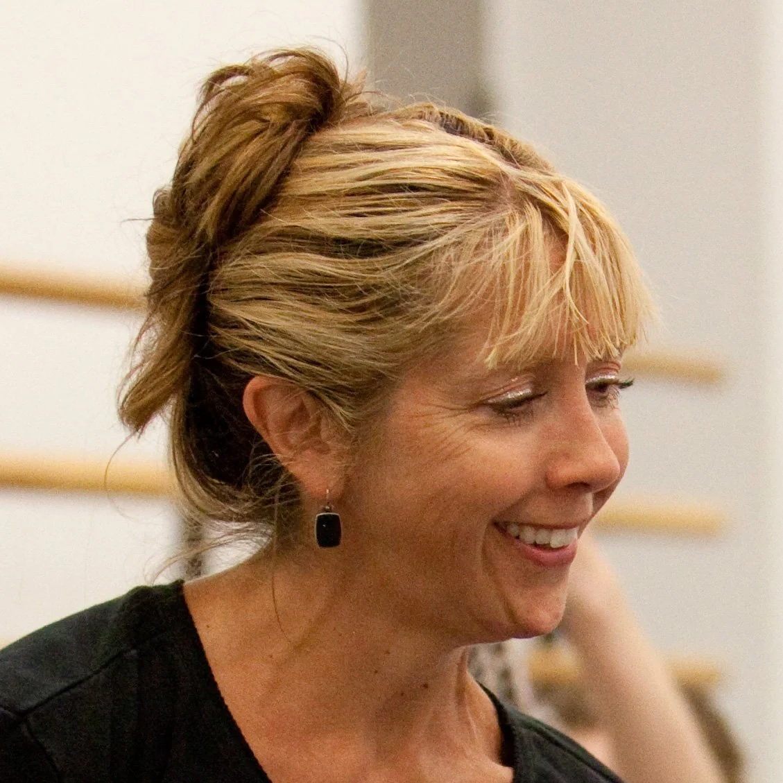 Close-up of a smiling woman with blonde hair styled in an updo, wearing earrings and a black top.