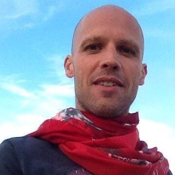 A man with a shaved head wearing a red bandana around his neck, outdoors with a cloudy sky in the background.