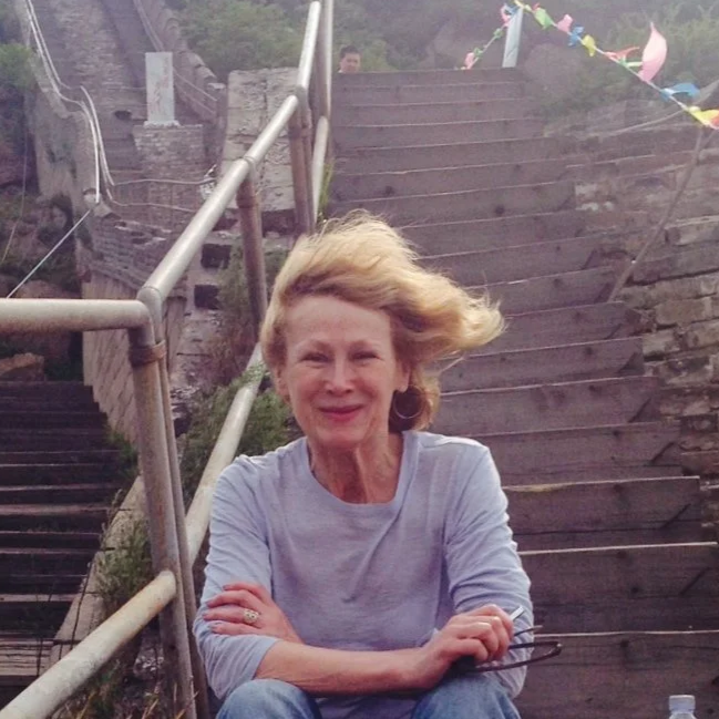 A smiling woman with shoulder-length blonde hair seated on the bottom of a staircase, with wind blowing her hair to the right. There are colorful decorations hanging in the background.