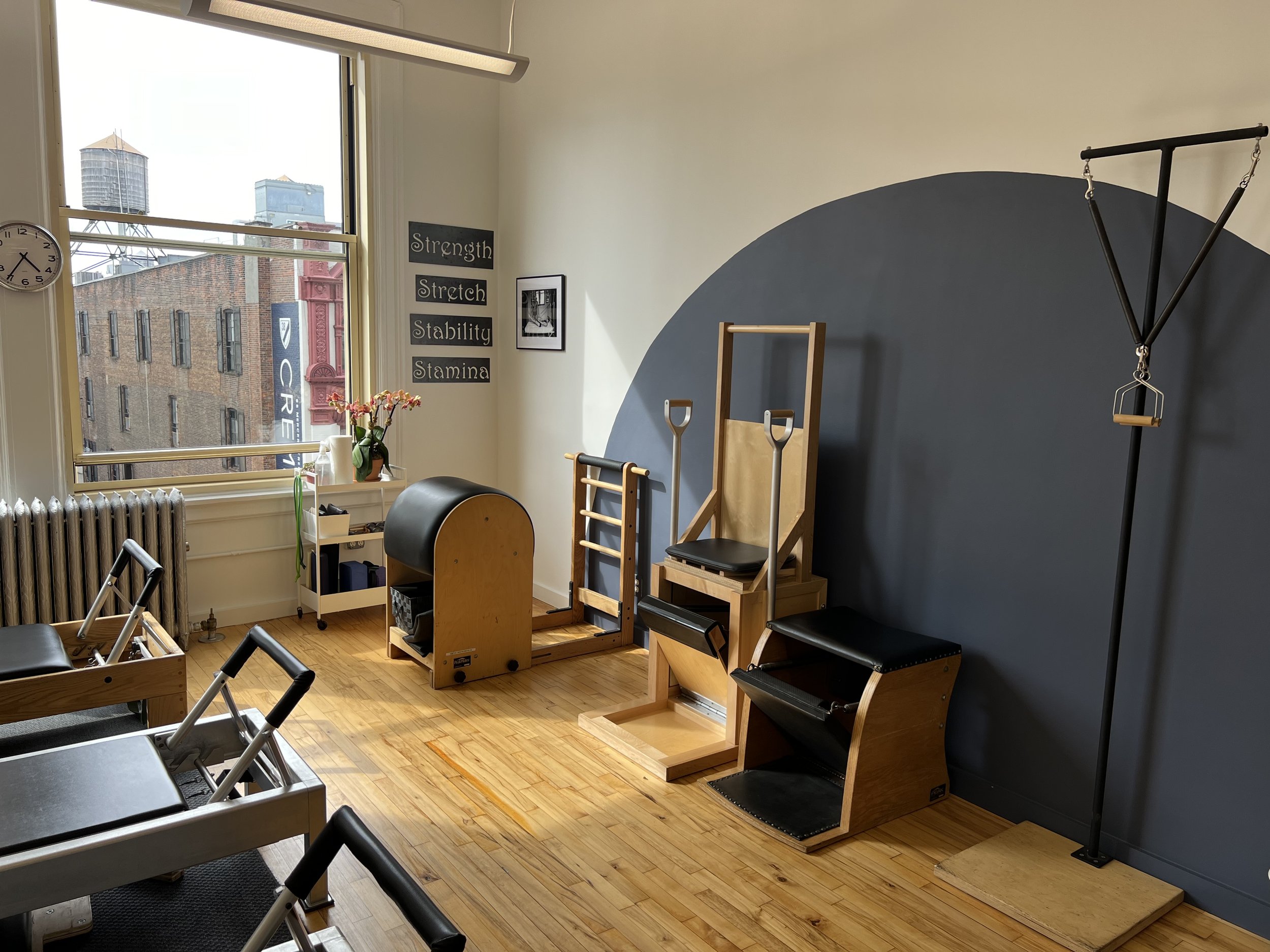 Interior of a fitness or Pilates studio with various workout equipment, a large window showing city buildings, and a dark blue accent wall with motivational words.