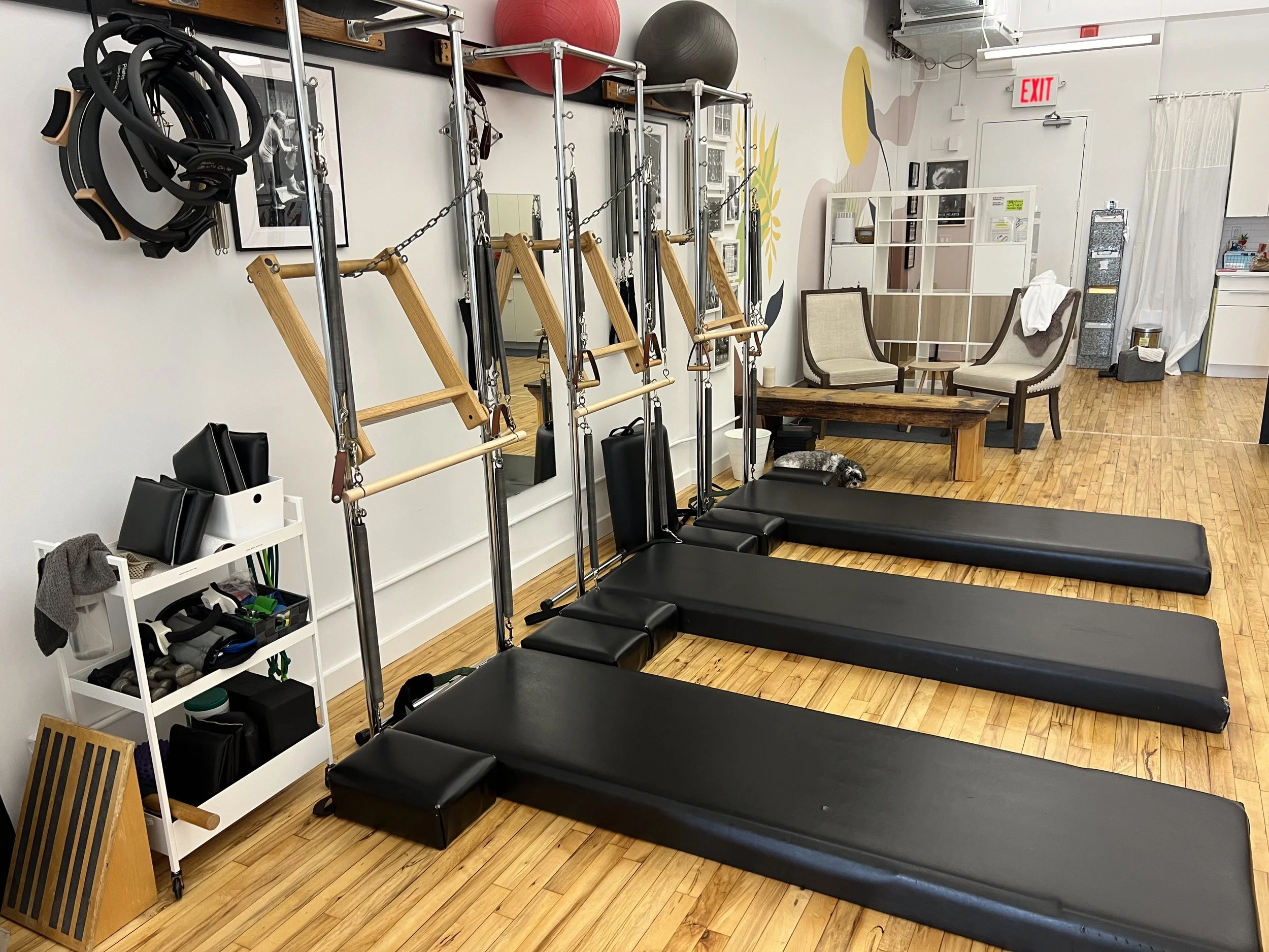 Pilates studio with three black padded mats, Pilates equipment, mirrors on the wall, and cozy seating area with two chairs and a small table.