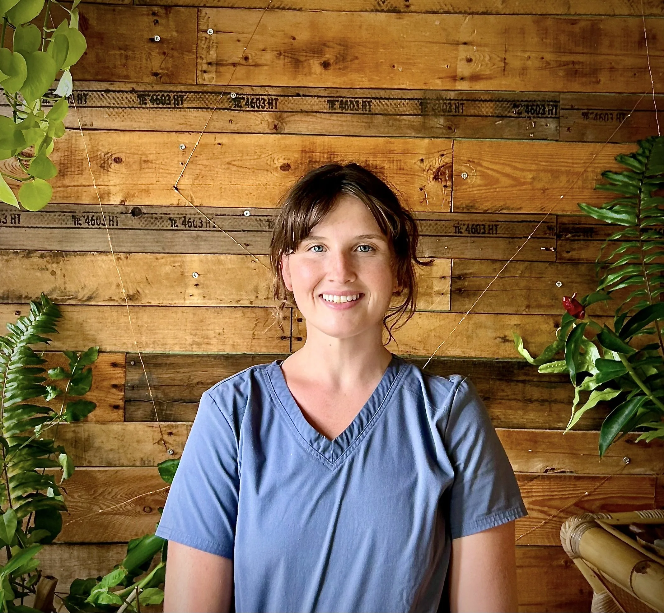 A smiling woman wearing blue scrubs sitting in front of a wooden wall with green plants on either side.