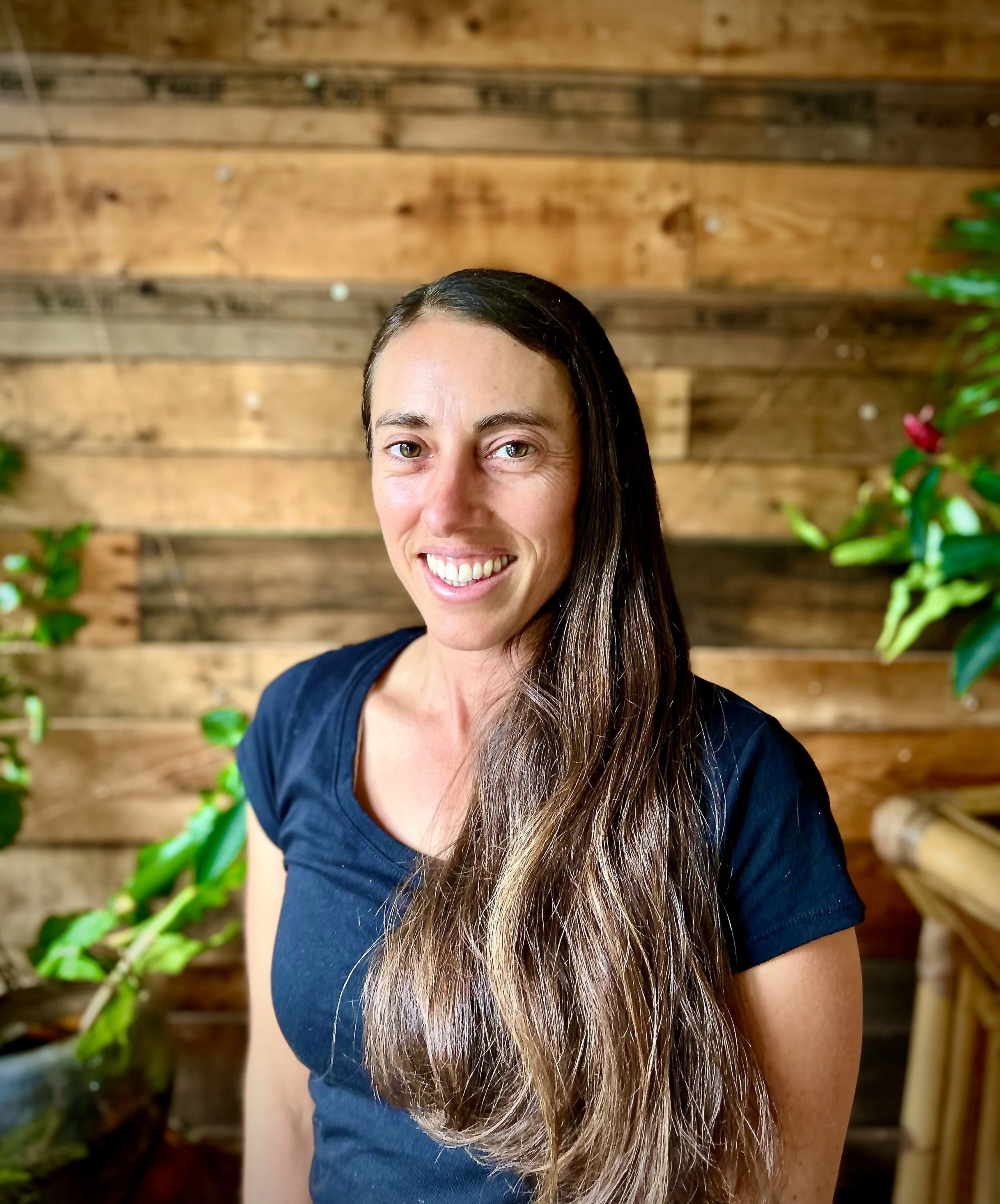 A woman with long brown hair wearing a black shirt, smiling, standing indoors in front of a wooden wall with green plants on the sides.