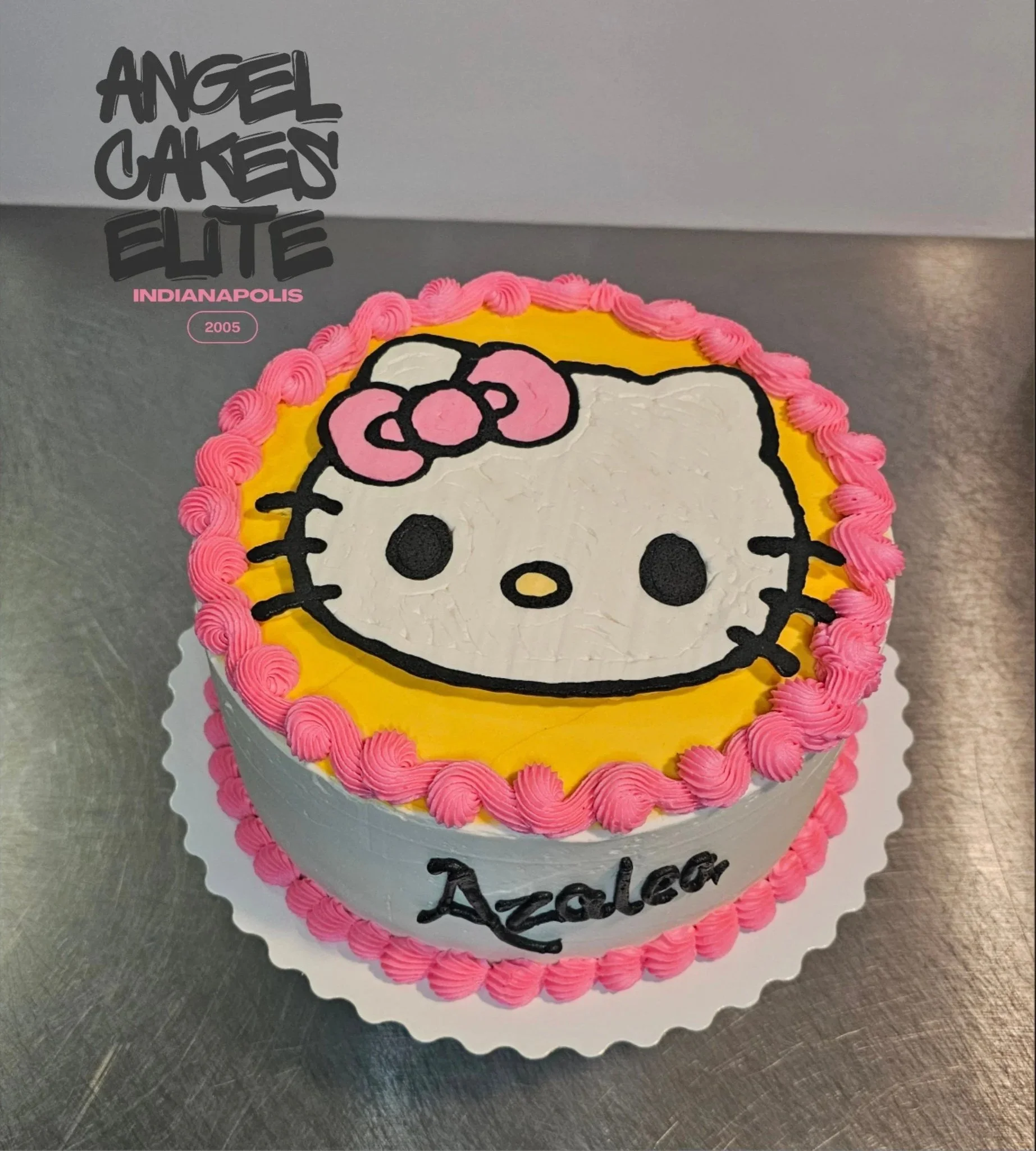 Cake featuring Hello Kitty with a pink bow, a yellow background, pink border, and the name 'Azaler' written on it.