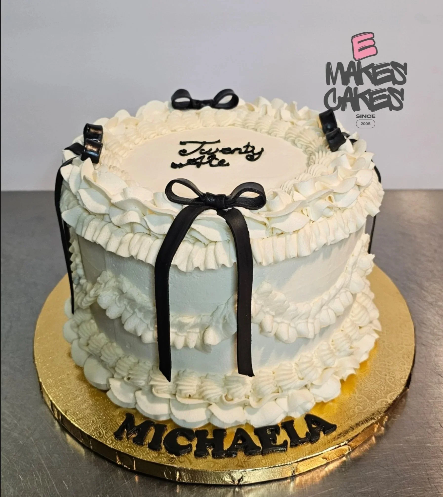 A white birthday cake decorated with black bows, with the words "Twenty one" written on top, and the name "Michaela" on the gold cake board, E Makes Cakes Indianapolis Cakes