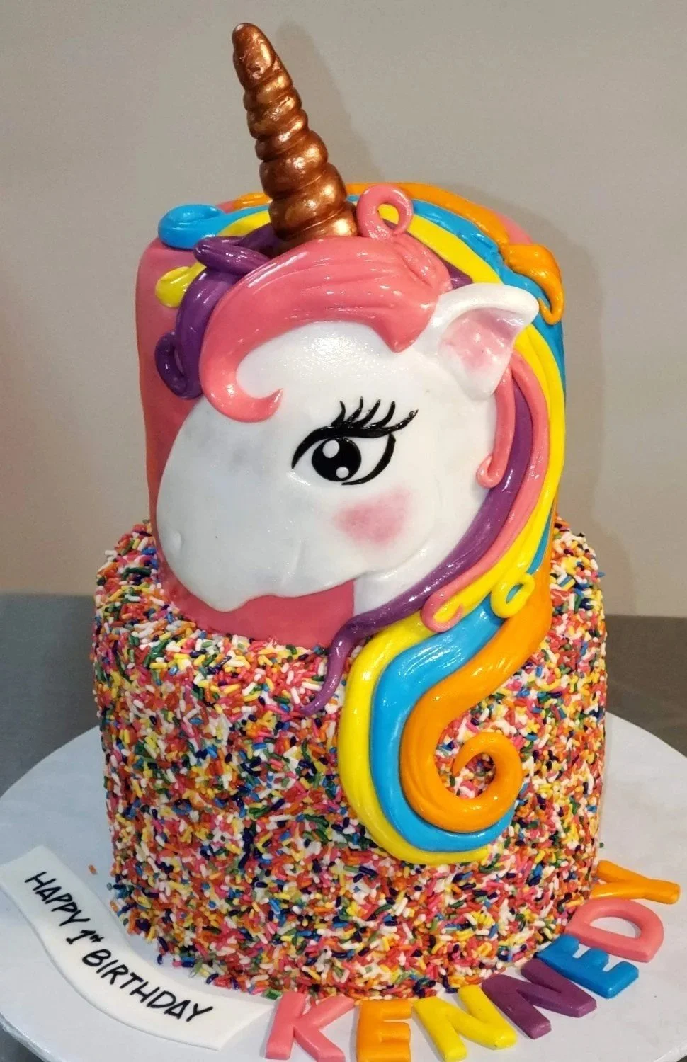Colorful unicorn-themed birthday cake with a rainbow mane, a gold horn, and sprinkles on the base, with vibrant letters spelling 'KENNEDY' on the cake board, E Makes Cakes, Indianapolis Cakes
